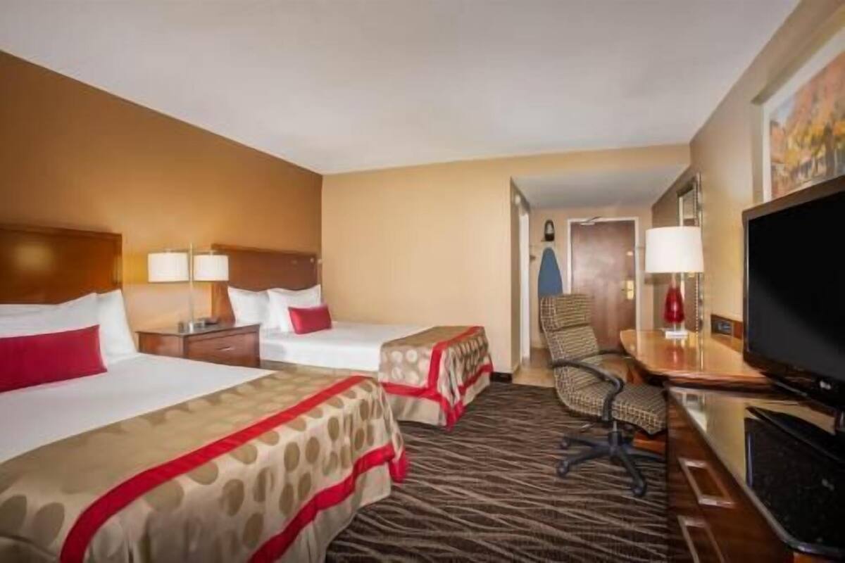 Habitación de hotel con dos camas, un escritorio con silla, televisor y una puerta que da a otra habitación.