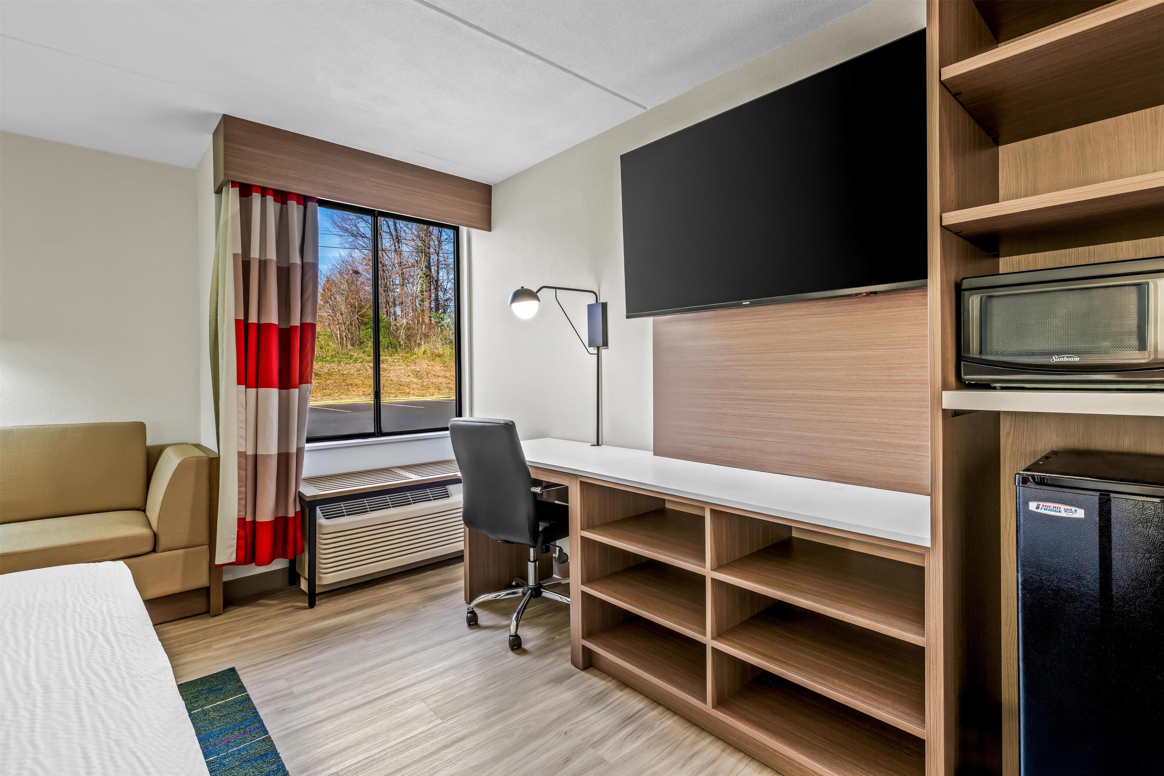 A modern hotel room with a bed, a desk, a chair, a television, and a built-in microwave.
