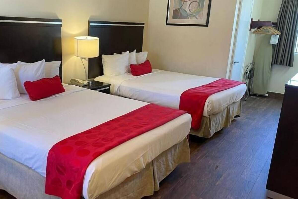 Duas camas de casal com roupa de cama branca e detalhes vermelhos, uma luminária de cabeceira e um quadro na parede.