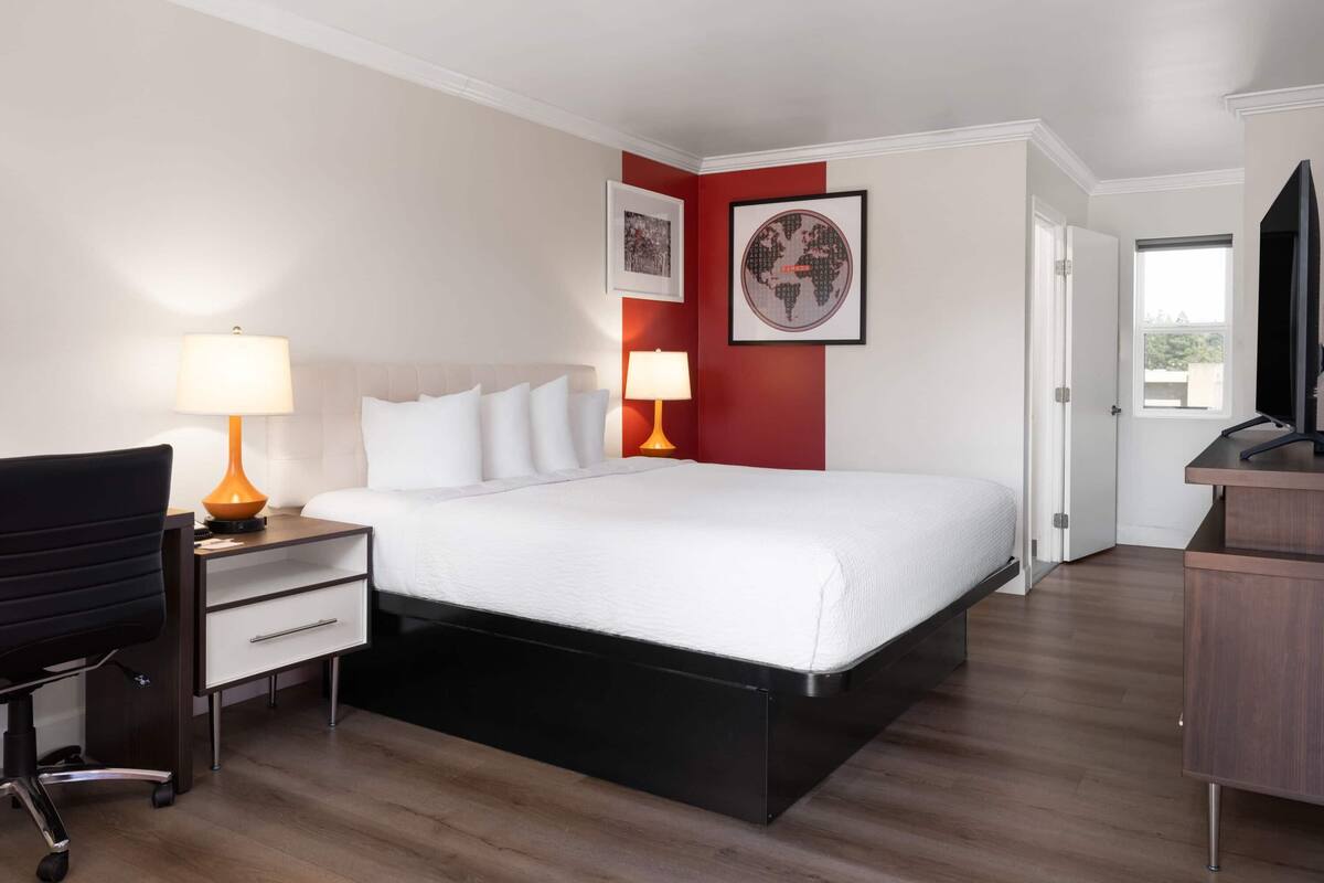 Quarto de hotel com cama, duas luminárias de cabeceira, uma escrivaninha com cadeira, televisão e porta.
