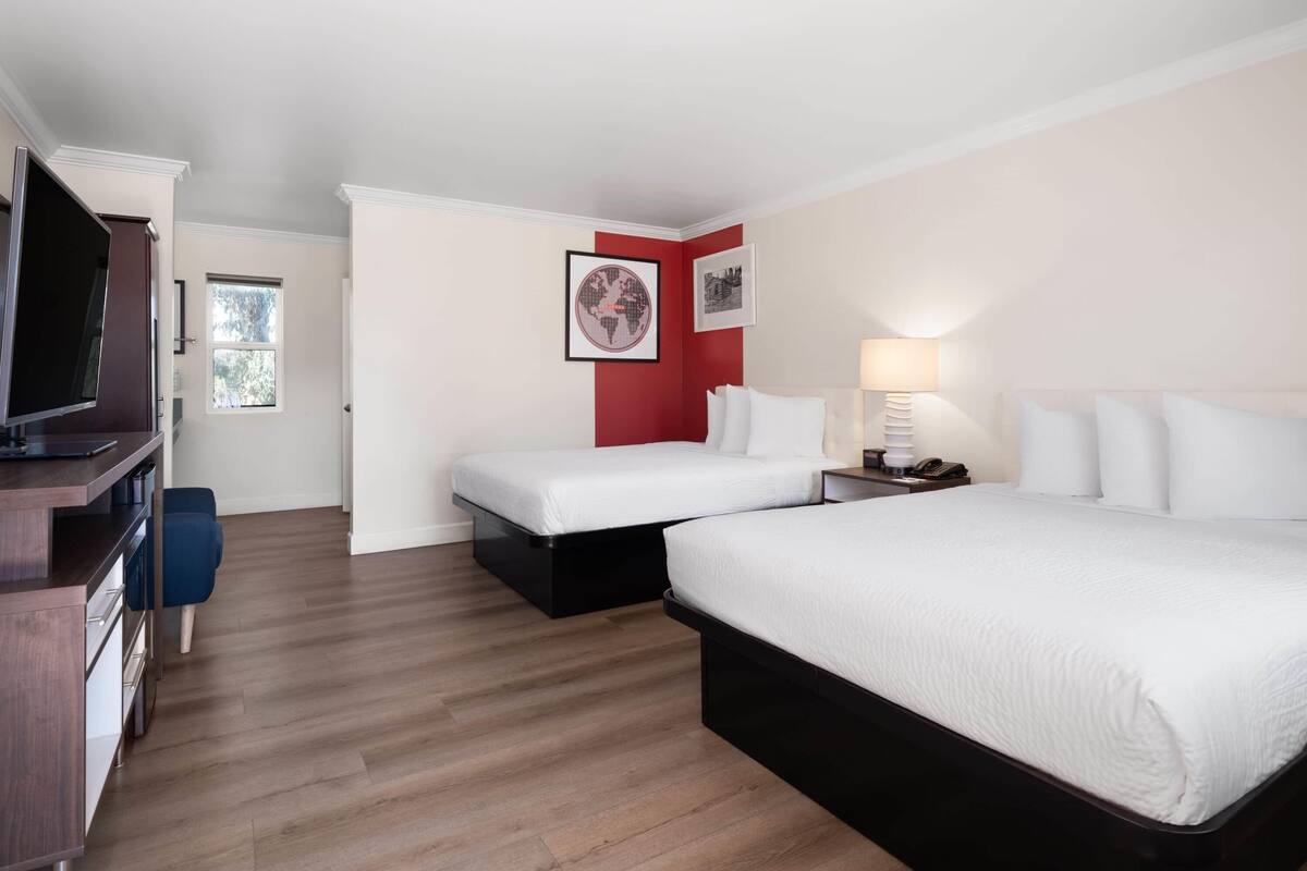Quarto de hotel com uma cama grande, uma cama menor, uma mesa com televisão e uma parede de destaque vermelha.