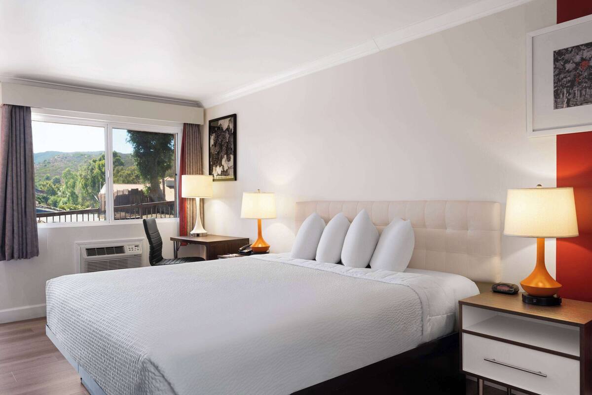 Quarto de hotel com uma cama grande, duas mesas de cabeceira com abajures, uma cadeira, uma escrivaninha e uma janela com vista para o verde.
