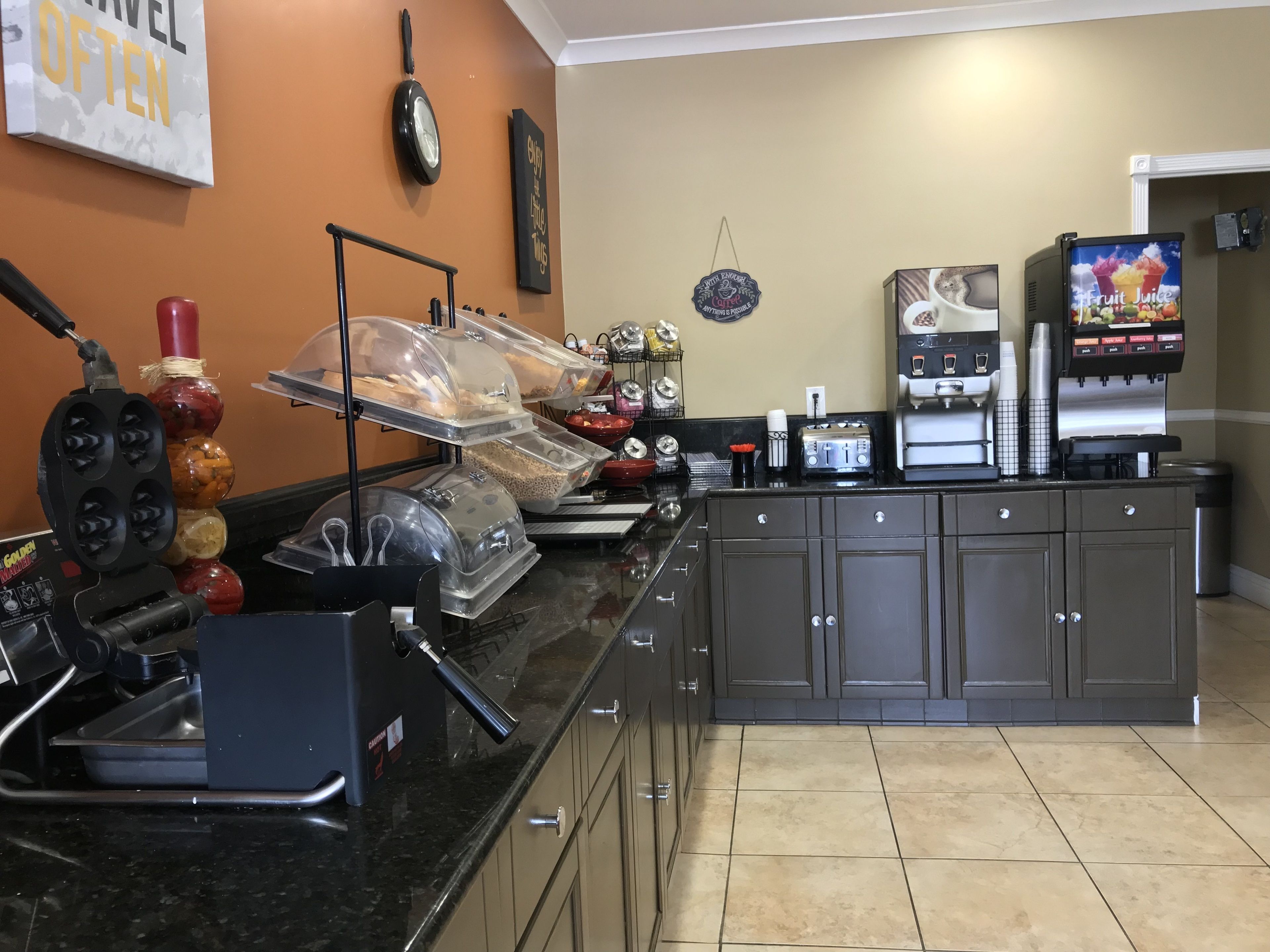 A self-service breakfast area with a waffle maker, coffee machine, and fruit juice dispenser.