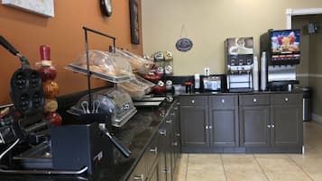 A self-service breakfast area with a waffle maker, coffee machine, and fruit juice dispenser.