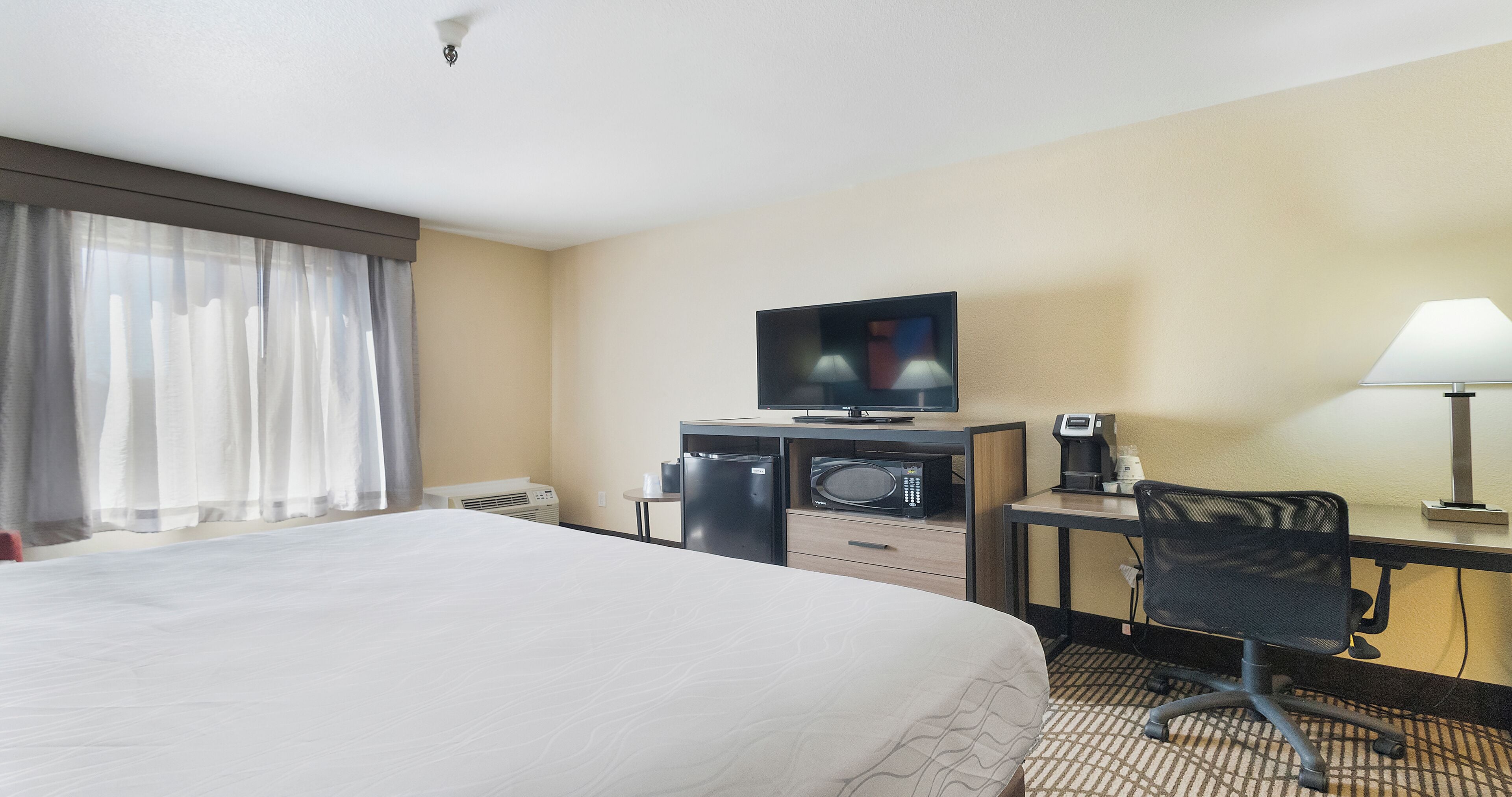 A hotel room with a bed, a desk with a computer, a microwave, a coffee maker, and a TV.