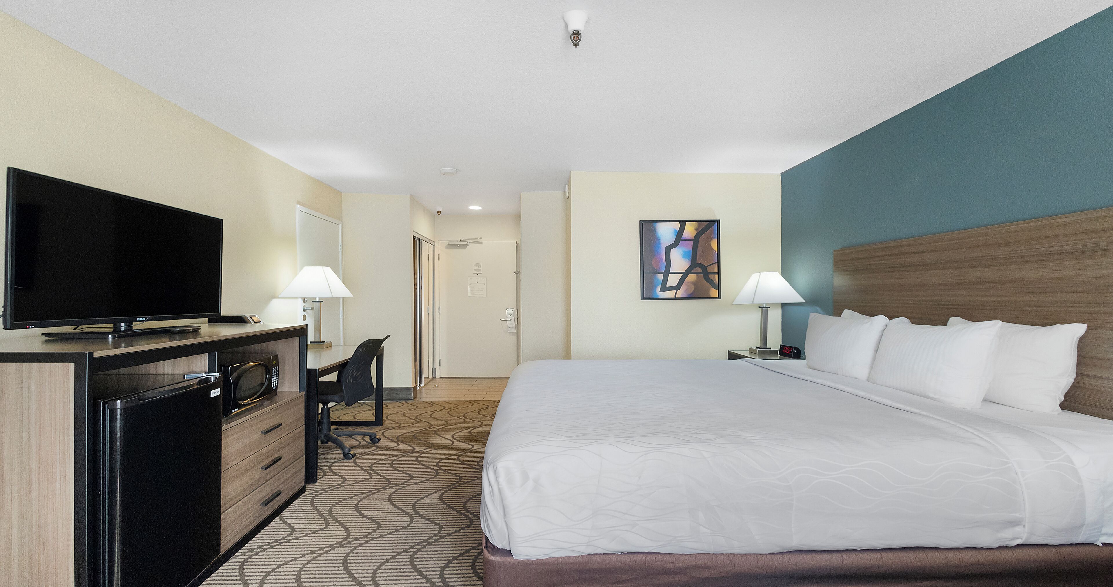 A hotel room with a bed, desk, television, and a painting on the wall.