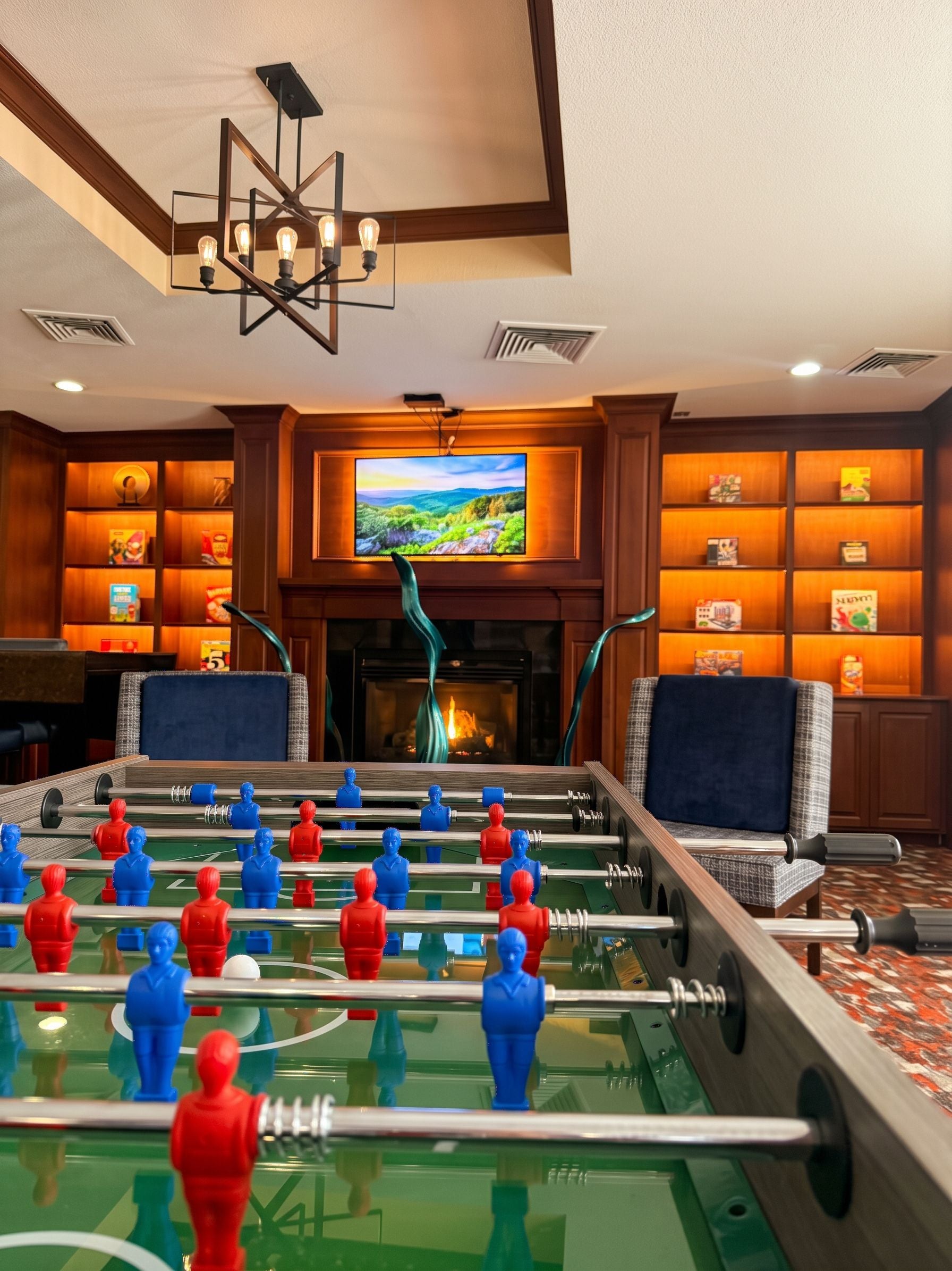 A room with a foosball table, a television mounted above a fireplace, and bookshelves.