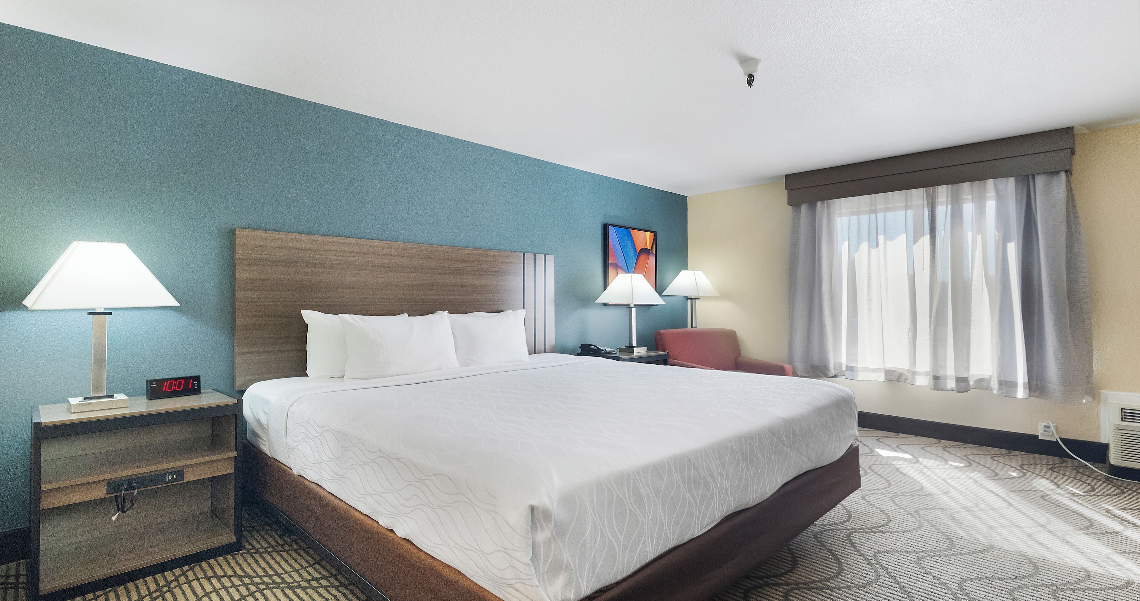 A hotel room with a large bed, two bedside tables with lamps, a chair, a window with curtains, and a wall clock.