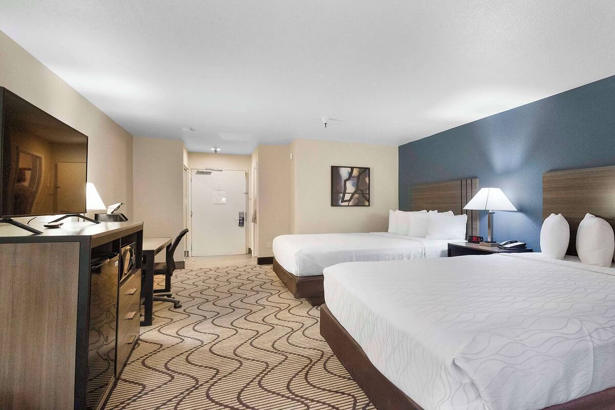 A hotel room with two beds, a desk, a chair, a nightstand, and a wall-mounted artwork.