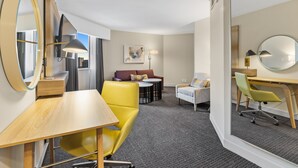 Ein modernes Hotelzimmer mit einem Holztisch, einem gelben Sessel und einer Couch mit Kissen. An der Wand hängt ein runder Spiegel und über der Couch ein Gemälde.