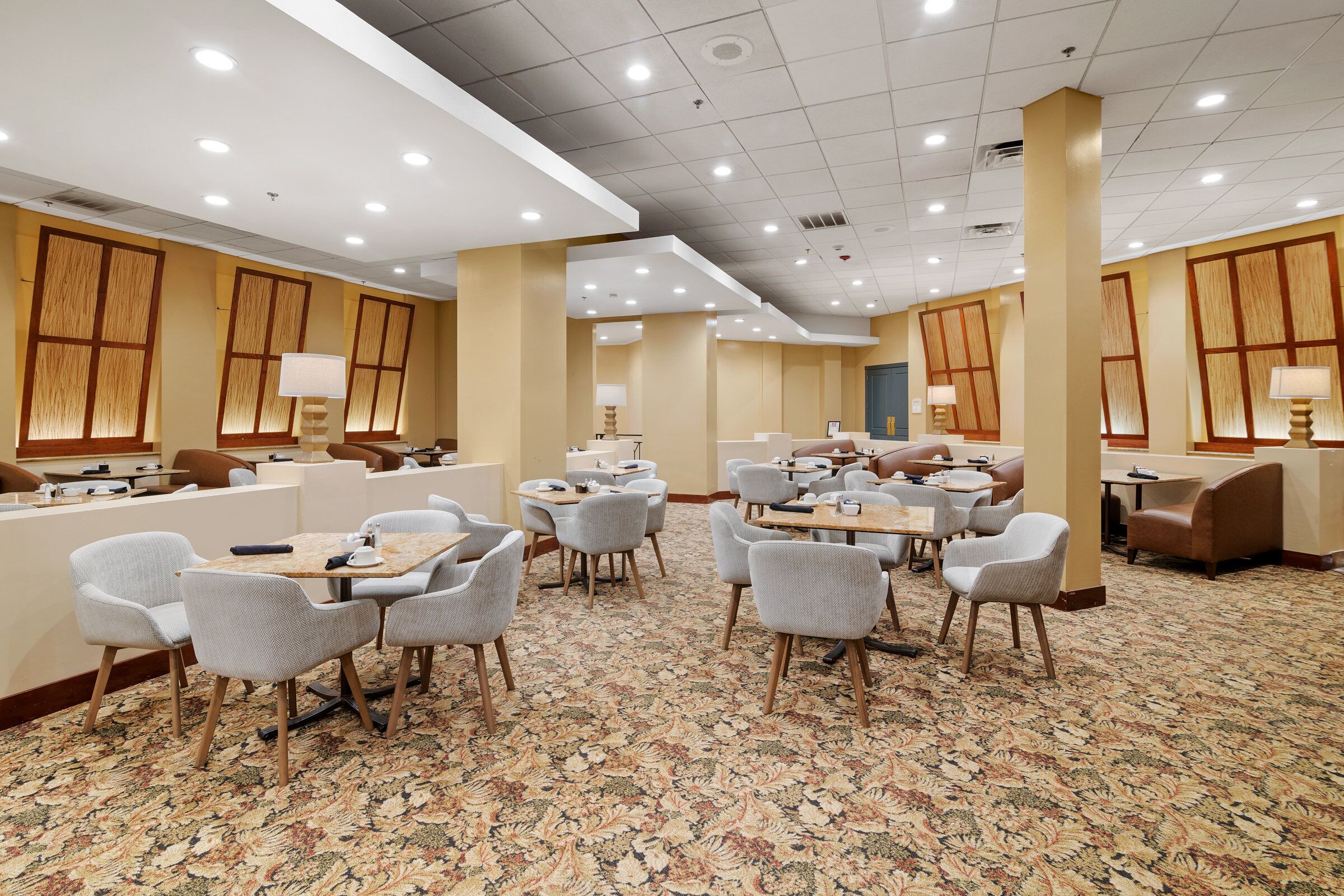 Um hall de hotel com várias áreas de estar, incluindo mesas redondas e cadeiras, e piso coberto por carpete.