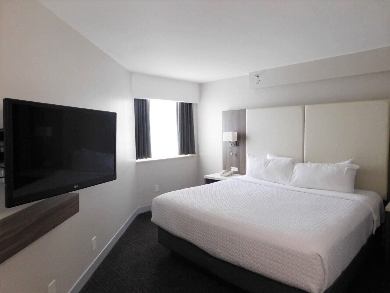 A hotel room with a large bed, a flat-screen TV mounted on the wall, a bedside table with a lamp, and a window with curtains.