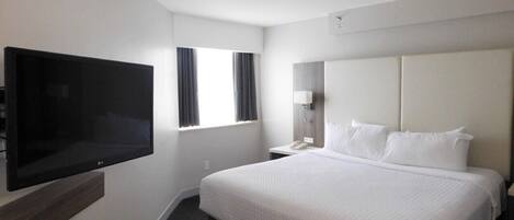 A hotel room with a large bed, a flat-screen TV mounted on the wall, a bedside table with a lamp, and a window with curtains.