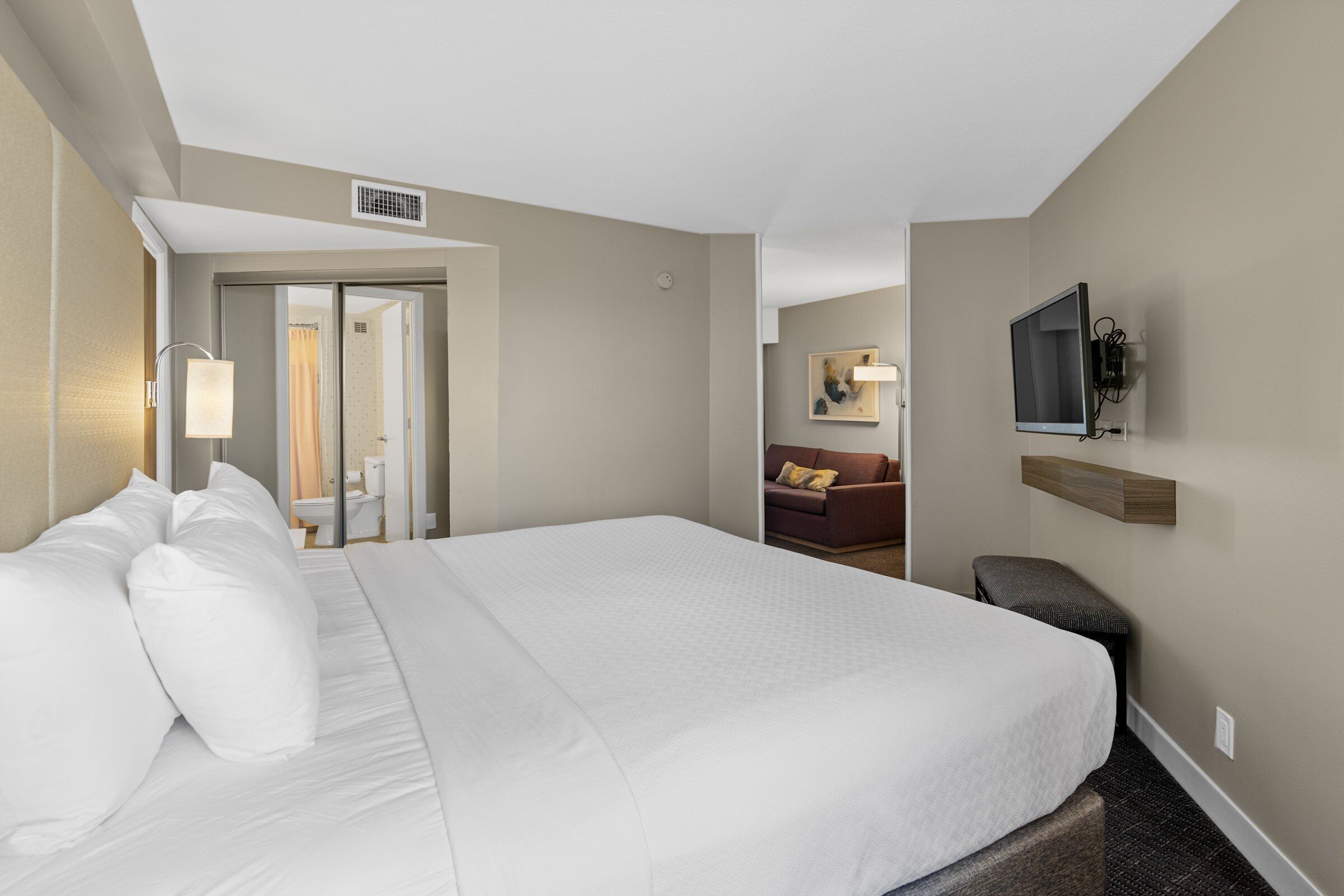 A hotel room with a large bed, a desk, a chair, a TV mounted on the wall, and a bathroom visible through an open door.