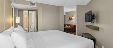 A hotel room with a large bed, a desk, a chair, a TV mounted on the wall, and a bathroom visible through an open door.