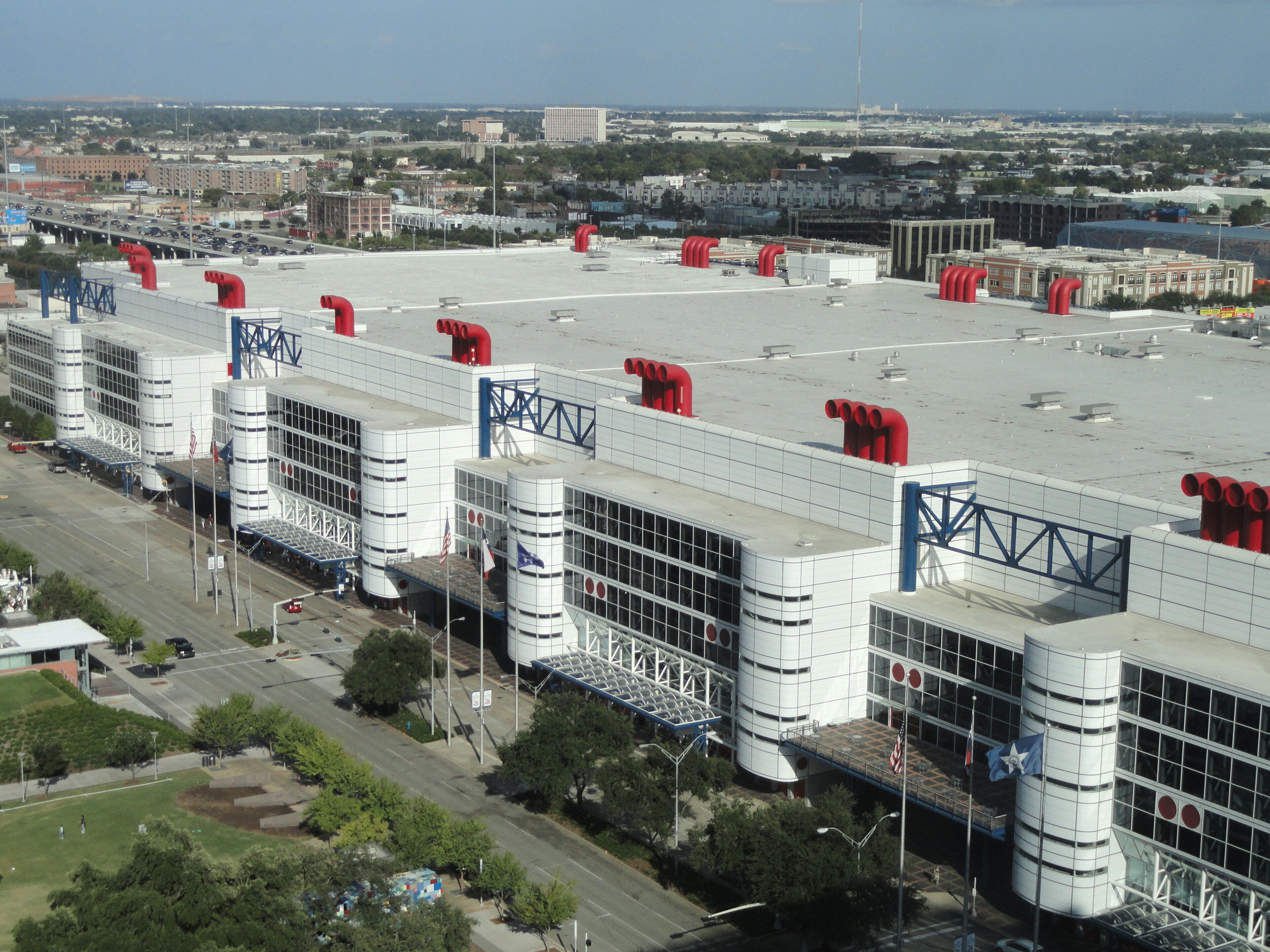 Un edificio industrial grande con varias estructuras cilíndricas y tuberías rojas, rodeado por un estacionamiento y una carretera.