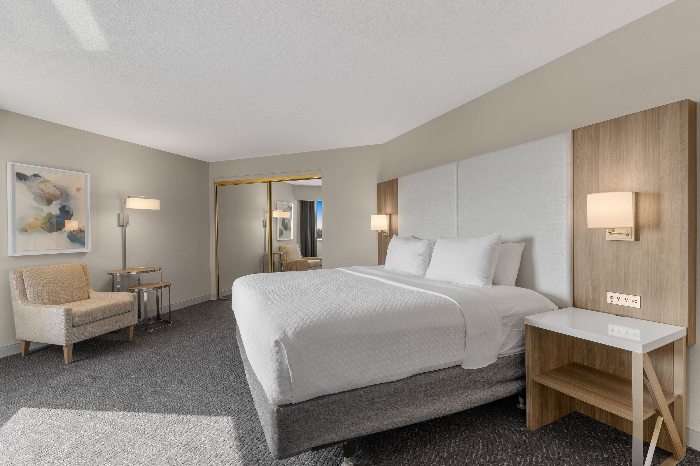 A modern hotel room with a large bed, a desk, a chair, and a small table.
