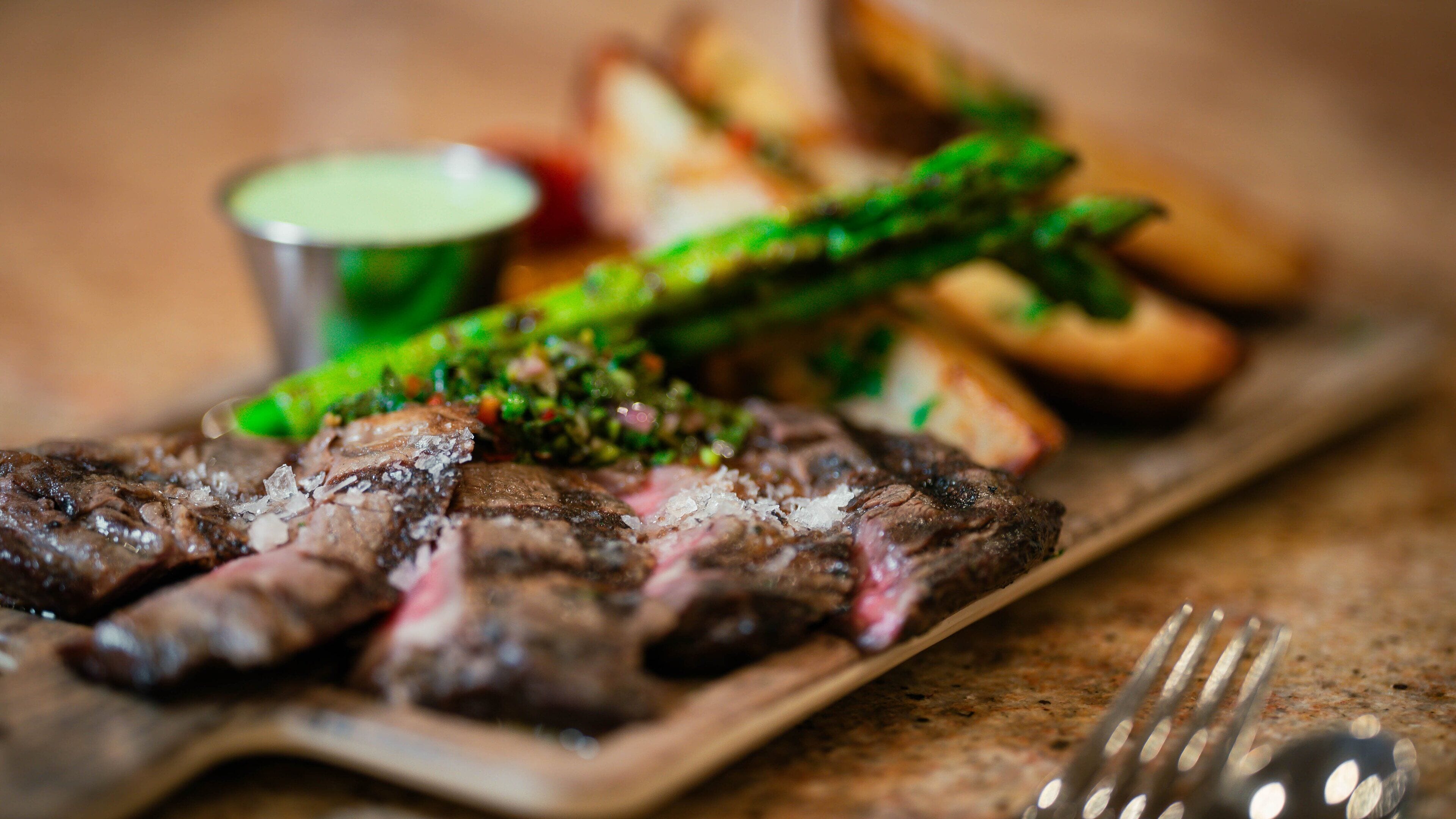 A plate of steak with asparagus and potatoes, accompanied by a small bowl of sauce.