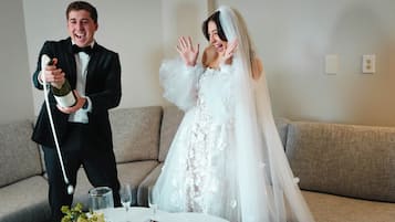 A wedding scene with a man in a tuxedo holding a bottle, a woman in a wedding dress, and a table with glasses and a flower arrangement.