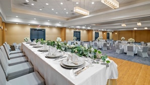 Ein Festsaal mit langen Tischen, festlich für eine Veranstaltung gedeckt, mit weißen Tischdecken, Silberbesteck und floralen Mittelpunkten.