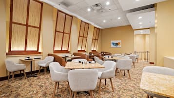 Un comedor de hotel con varias mesas y sillas, ventanas amplias y una alfombra estampada.