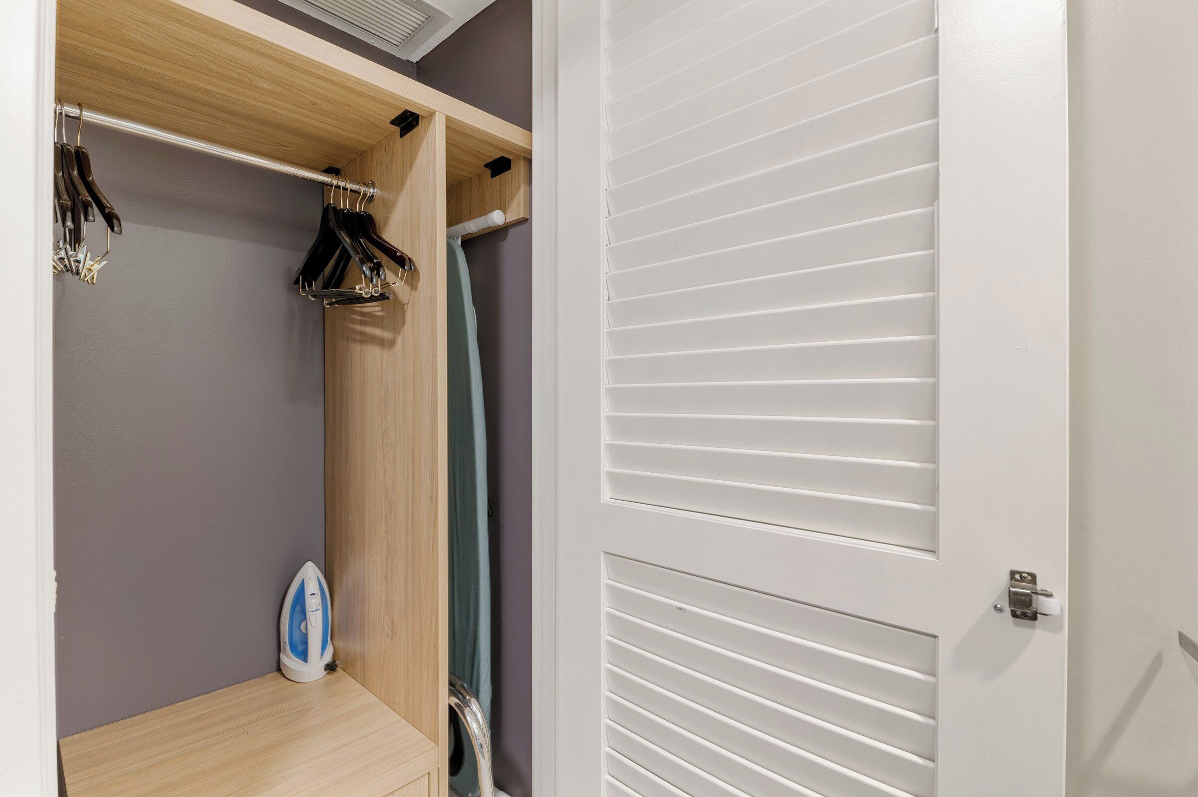 Une pièce avec une étagère en bois, un portant à vêtements suspendu et une porte blanche à lattes horizontales.