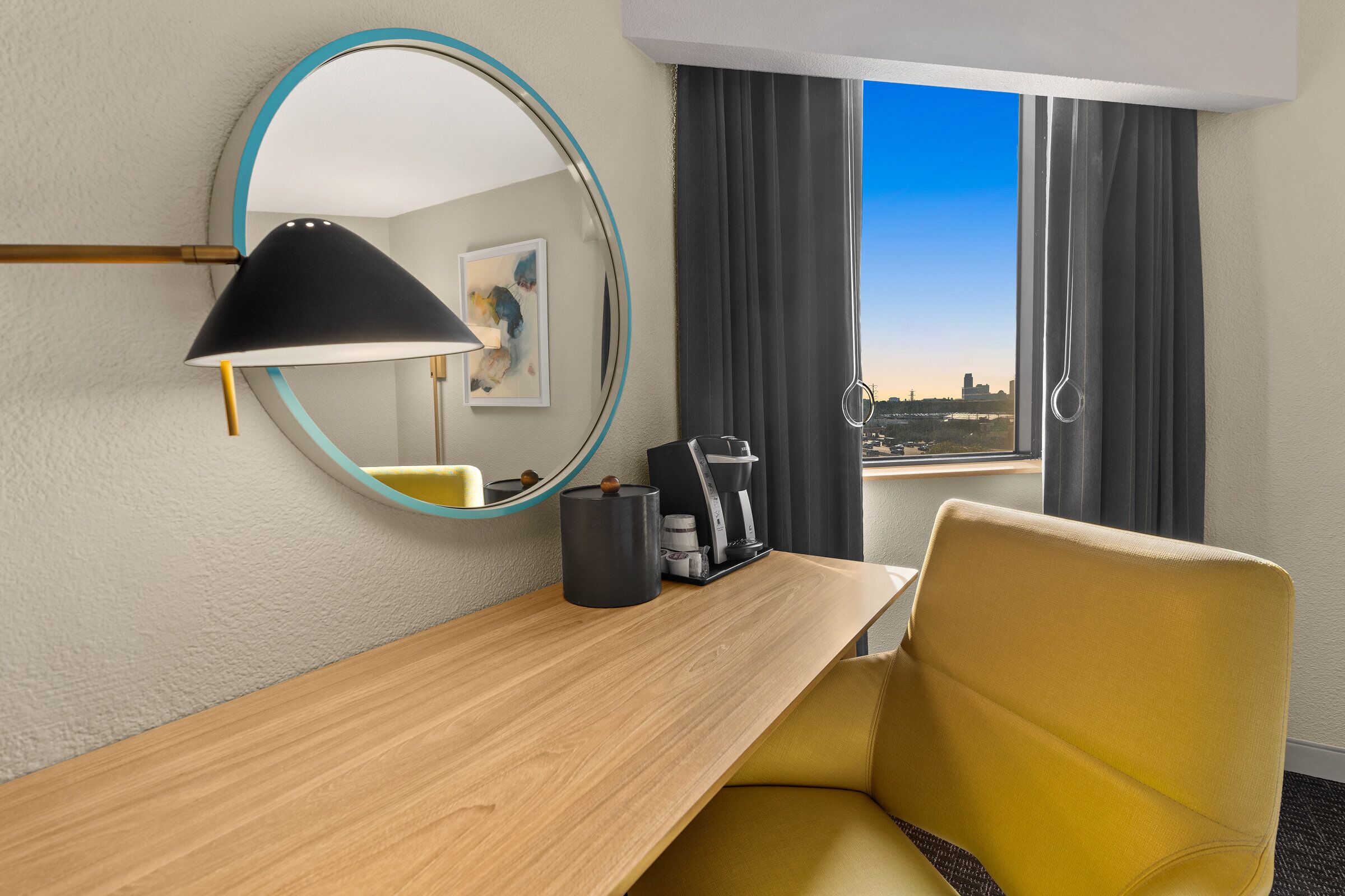 A hotel room with a wooden desk, a yellow chair, a black lamp, a coffee maker, and a view of the city through large windows.