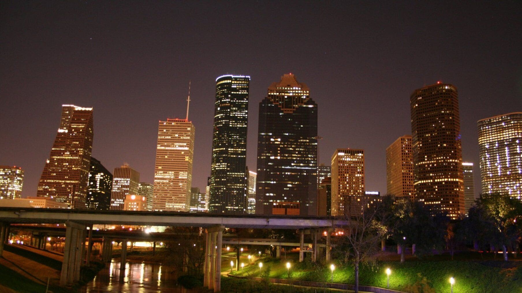 A nighttime cityscape with illuminated skyscrapers and a bridge.