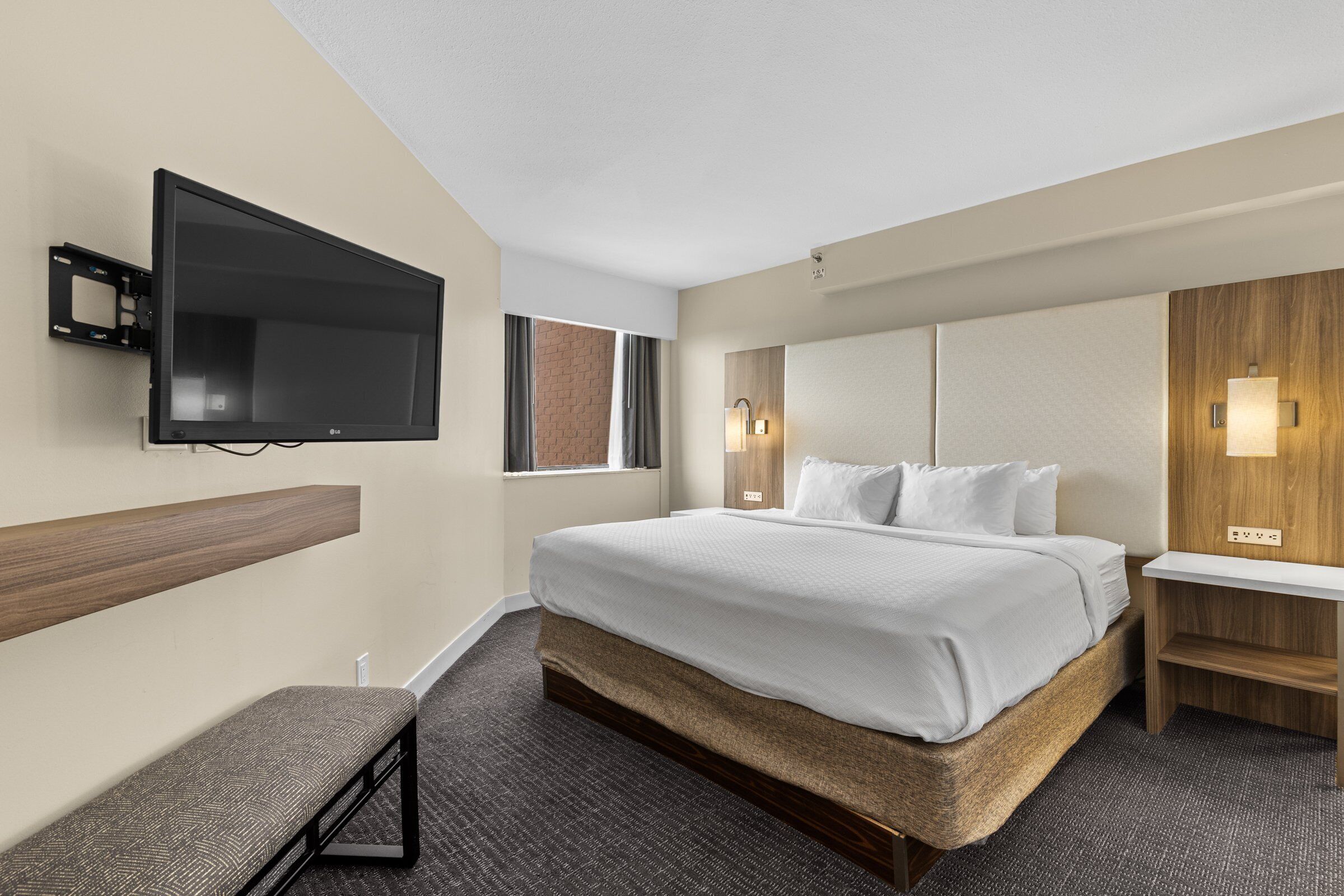 A hotel room with a large bed, a flat-screen TV mounted on the wall, a bench, and a small table.