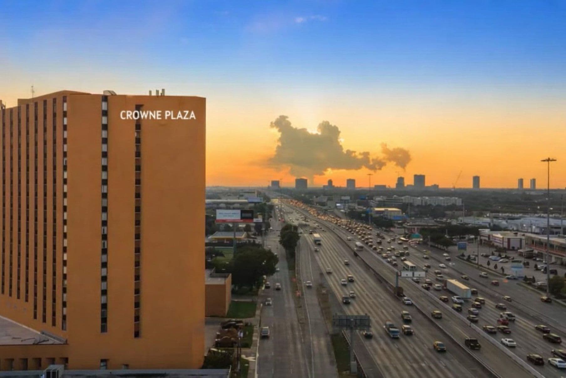 A multi-story building with the sign 'CROWNE PLAZA' on its facade, surrounded by a busy highway with multiple lanes of traffic.