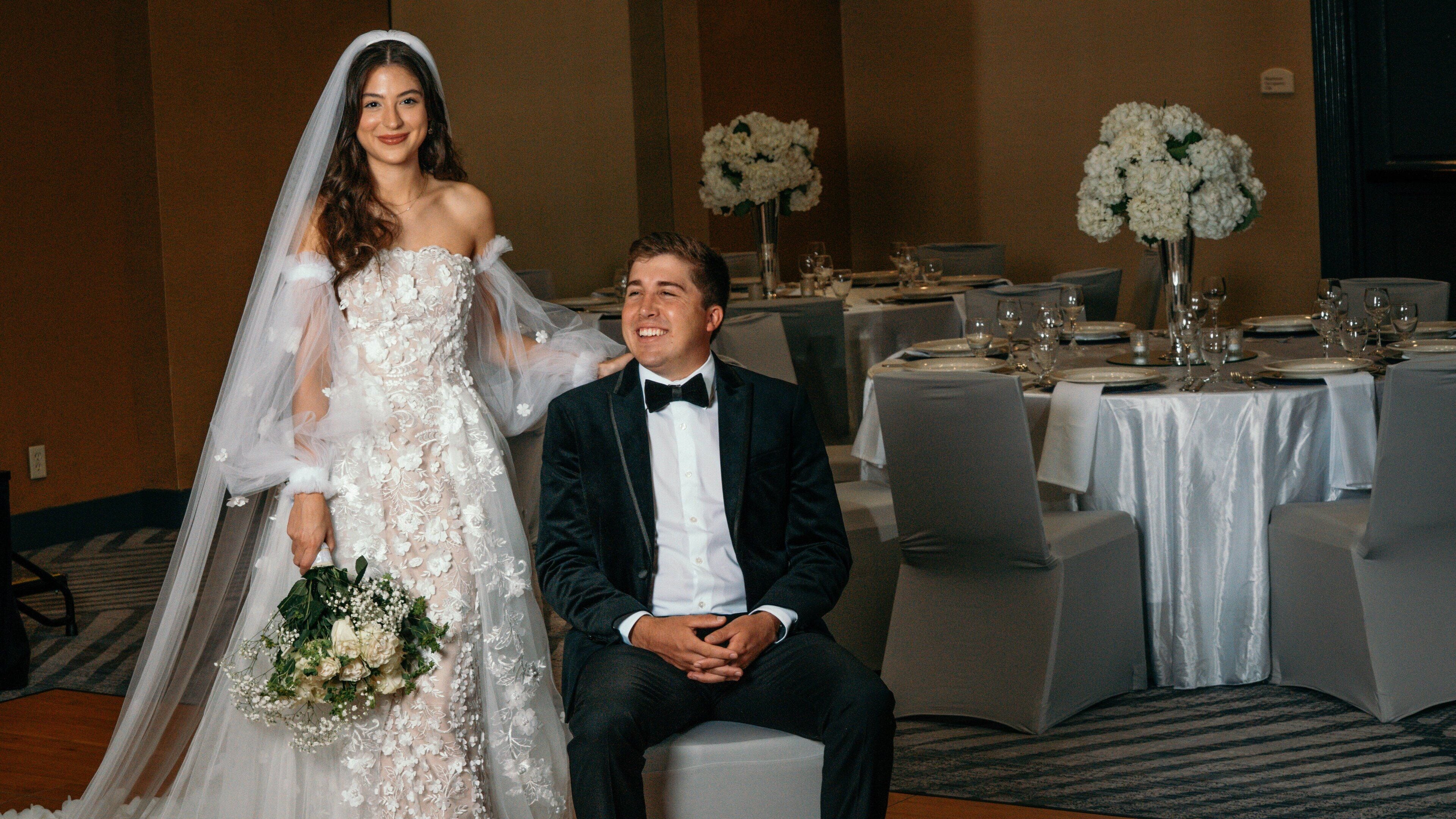 A wedding reception with a bride and groom seated at a table. Tables are covered with white tablecloths, and there are floral centerpieces on each.