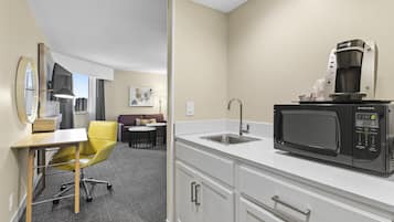 A compact hotel room with a kitchenette, including a microwave, coffee maker, and sink. There is a small dining area with a table and chairs, and a yellow office chair.