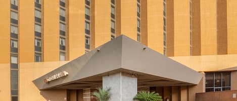 Entrada de hotel con diseño moderno, que presenta un pilar grande y un jardín circular con diversas plantas.
