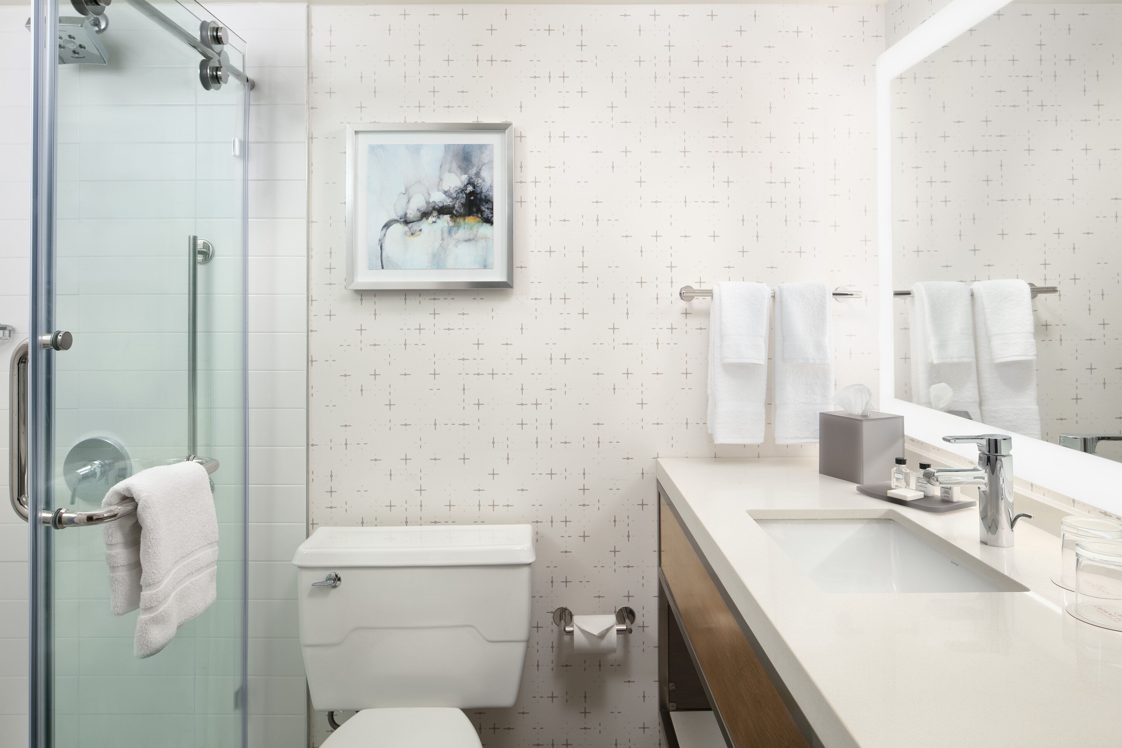 A modern bathroom with a glass shower enclosure, a toilet, a sink with a mirror, and a framed abstract painting on the wall.