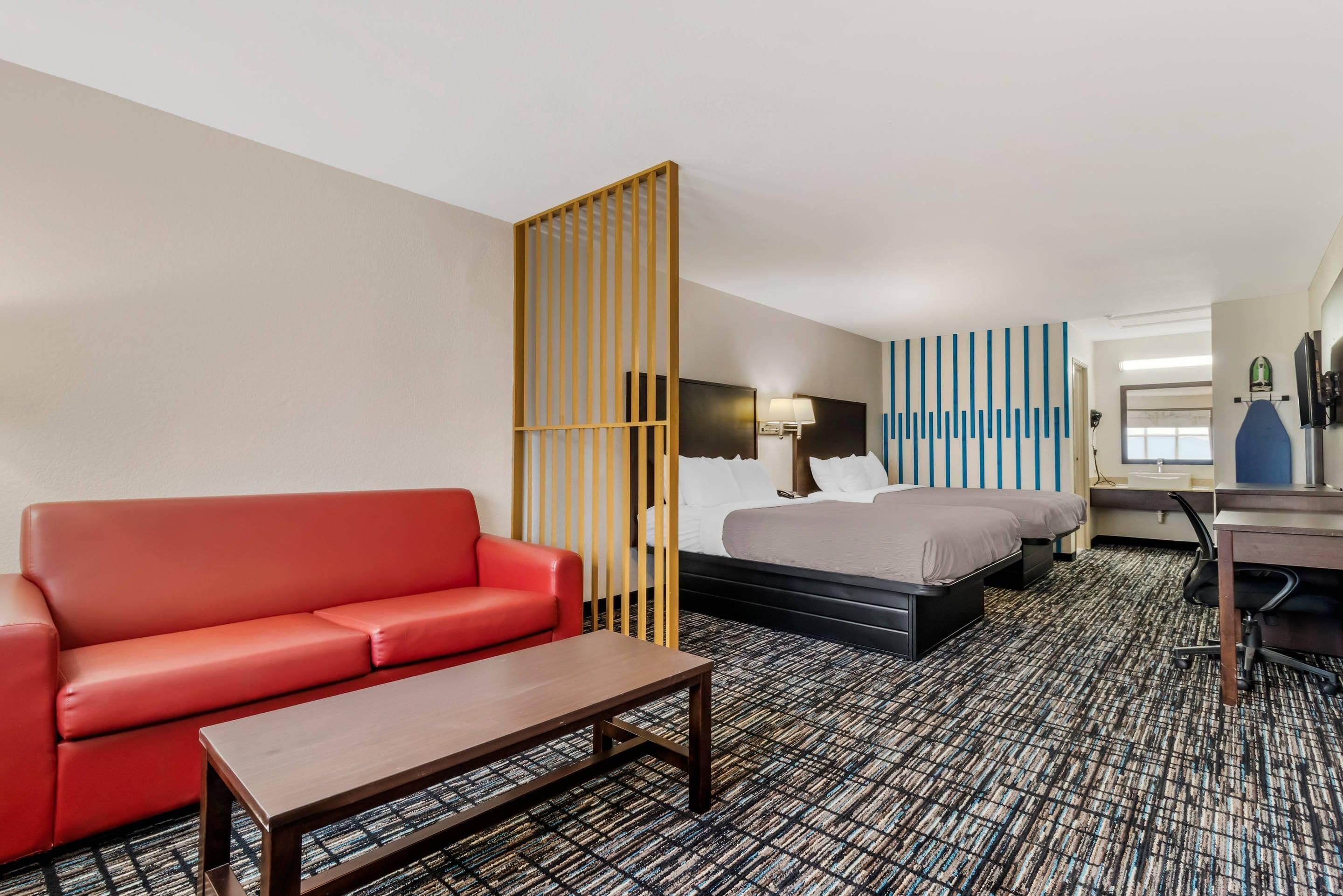 A hotel room with a red sofa, a bed with two pillows, a desk with a chair, and a patterned carpet.