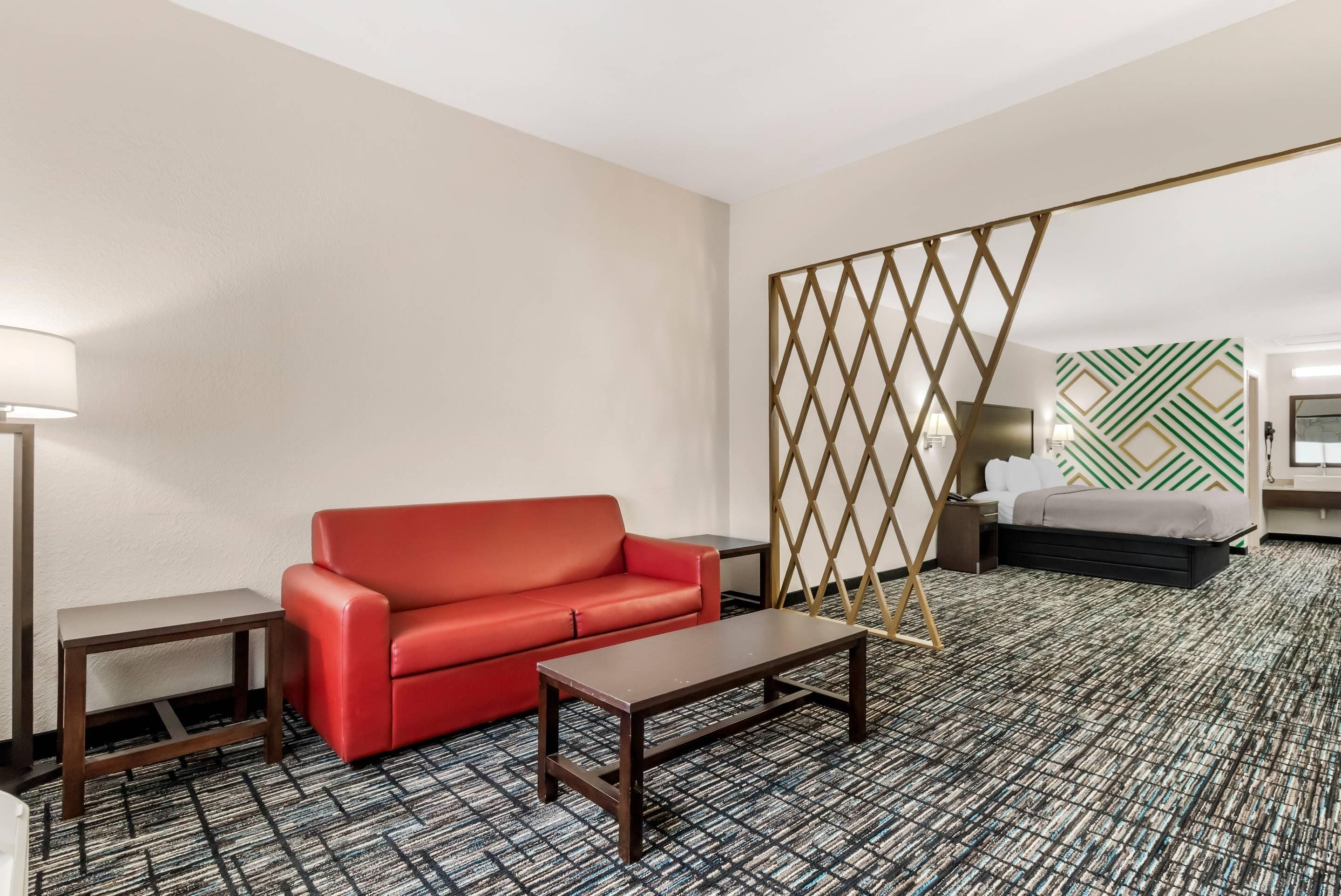 A hotel room with a red sofa, a small table, a decorative partition, a patterned carpet, and a bed with a green and white geometric wall design.