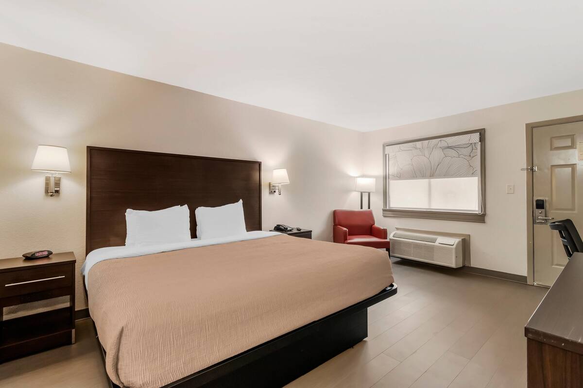 Una camera d'albergo con un letto grande, due lampade ai comodini, una poltrona rossa, una scrivania e una finestra vista.