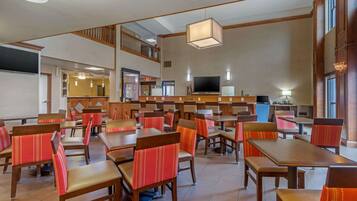 Een restaurantruimte in een hotel met meerdere tafels en stoelen, een televisie en een trap naar een hoger gelegen verdieping.