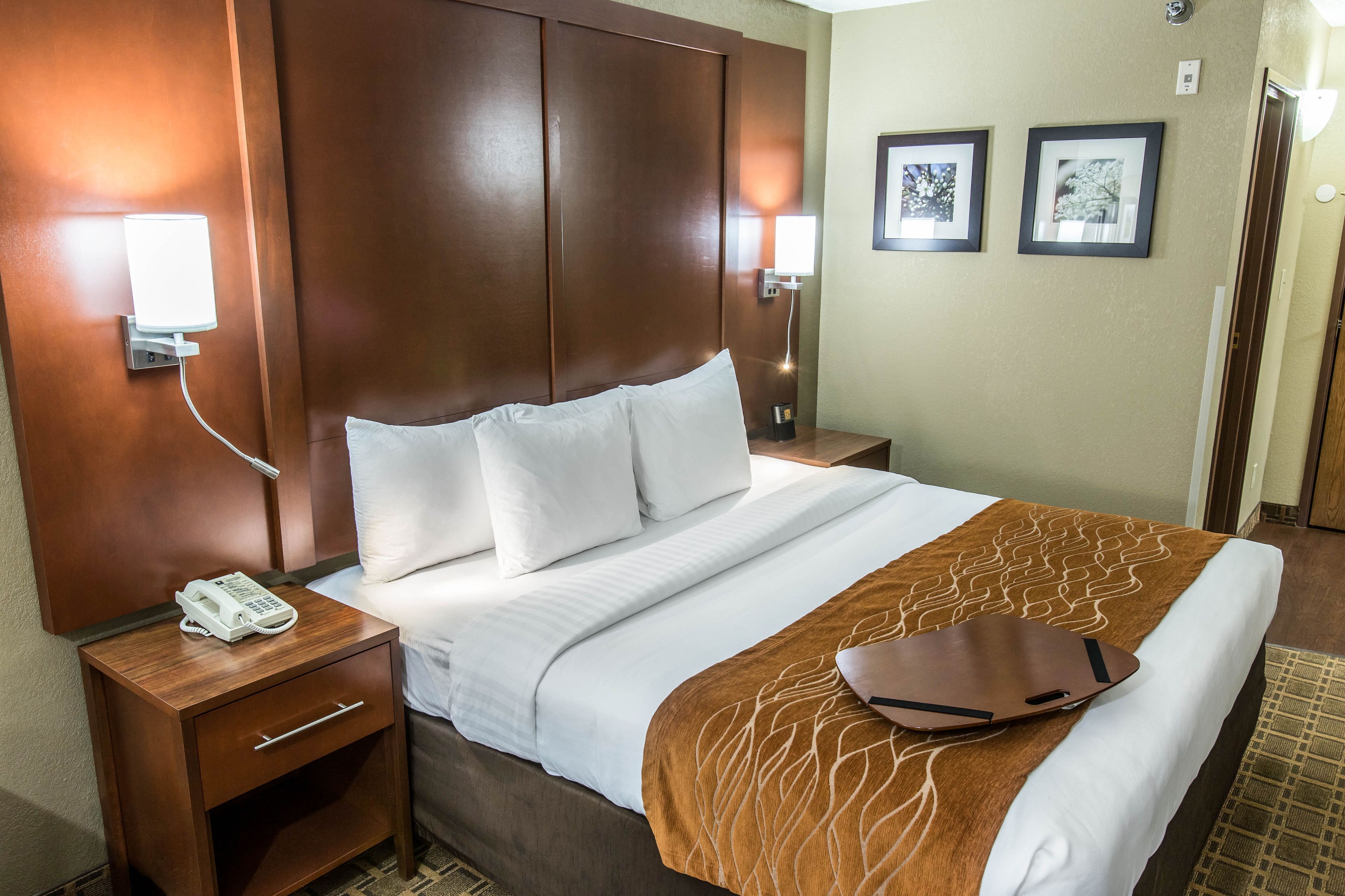 A hotel room with a large bed, a wooden nightstand with a phone, a brown throw blanket, and two framed pictures on the wall.