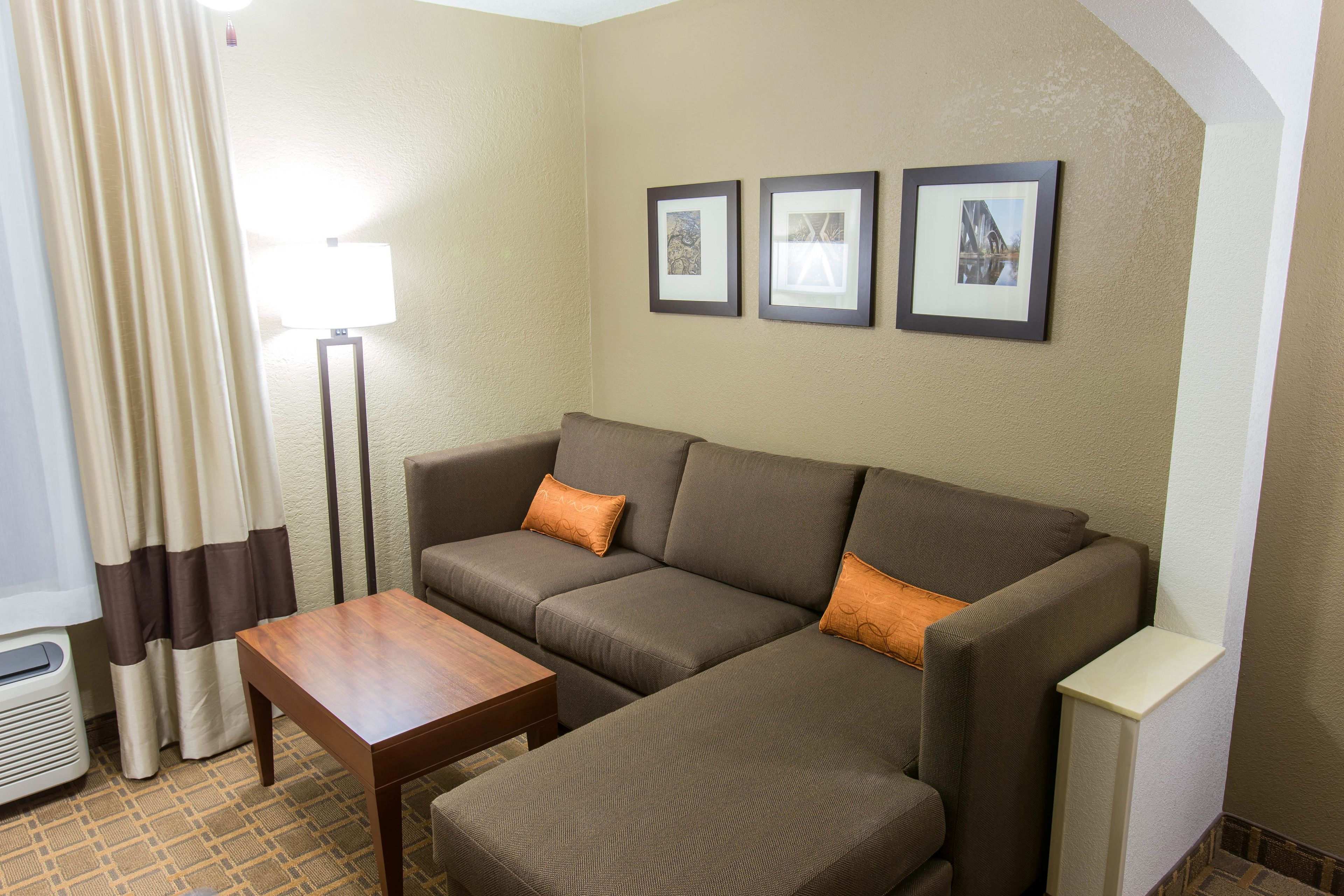 A living room with a brown sectional sofa, a wooden coffee table, a floor lamp, and a radiator. There are two framed pictures on the wall.