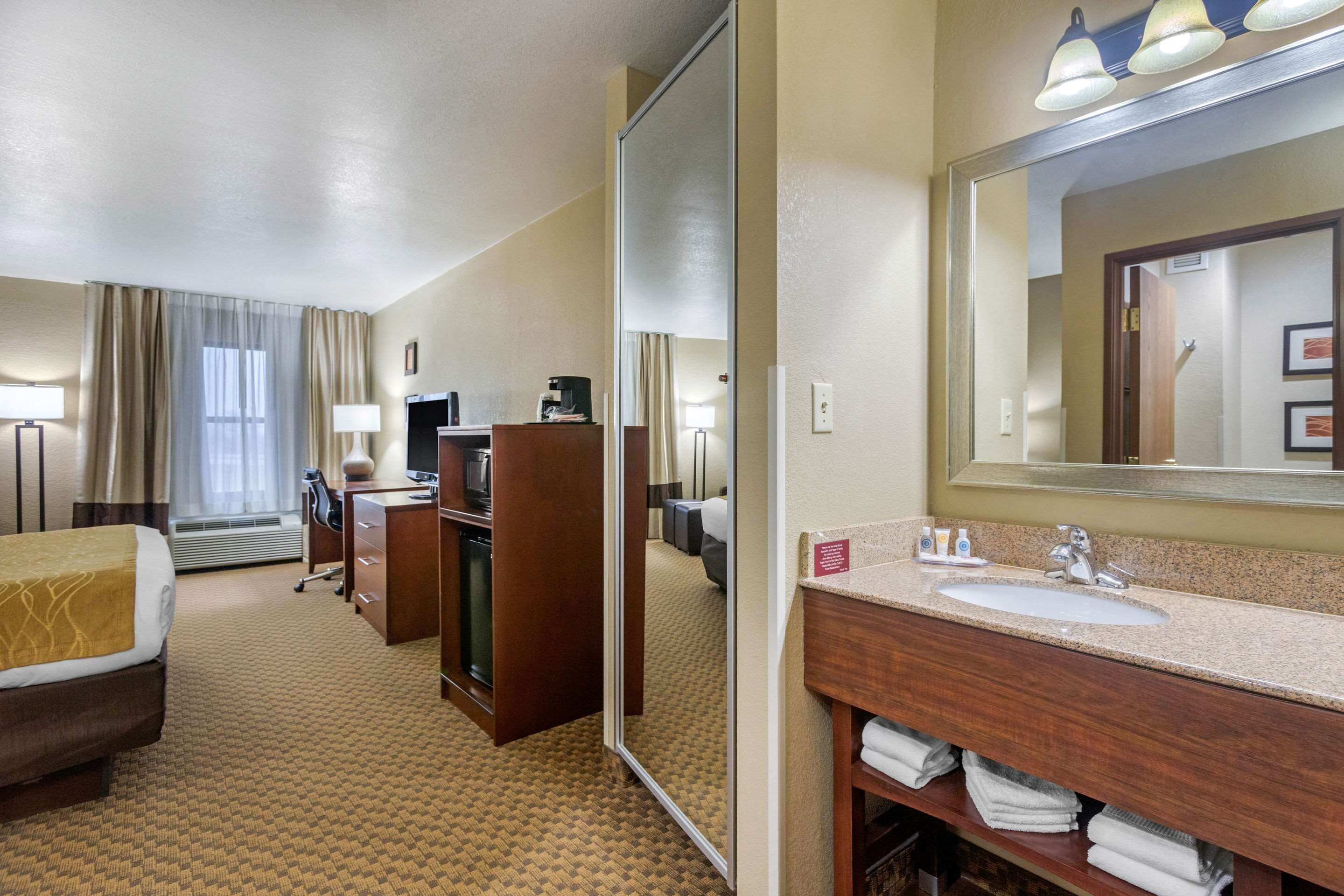 Une chambre d’hôtel comprenant un lit, un bureau, une chaise, une télévision, un miroir, un lavabo et une salle de bain avec un porte-serviettes.