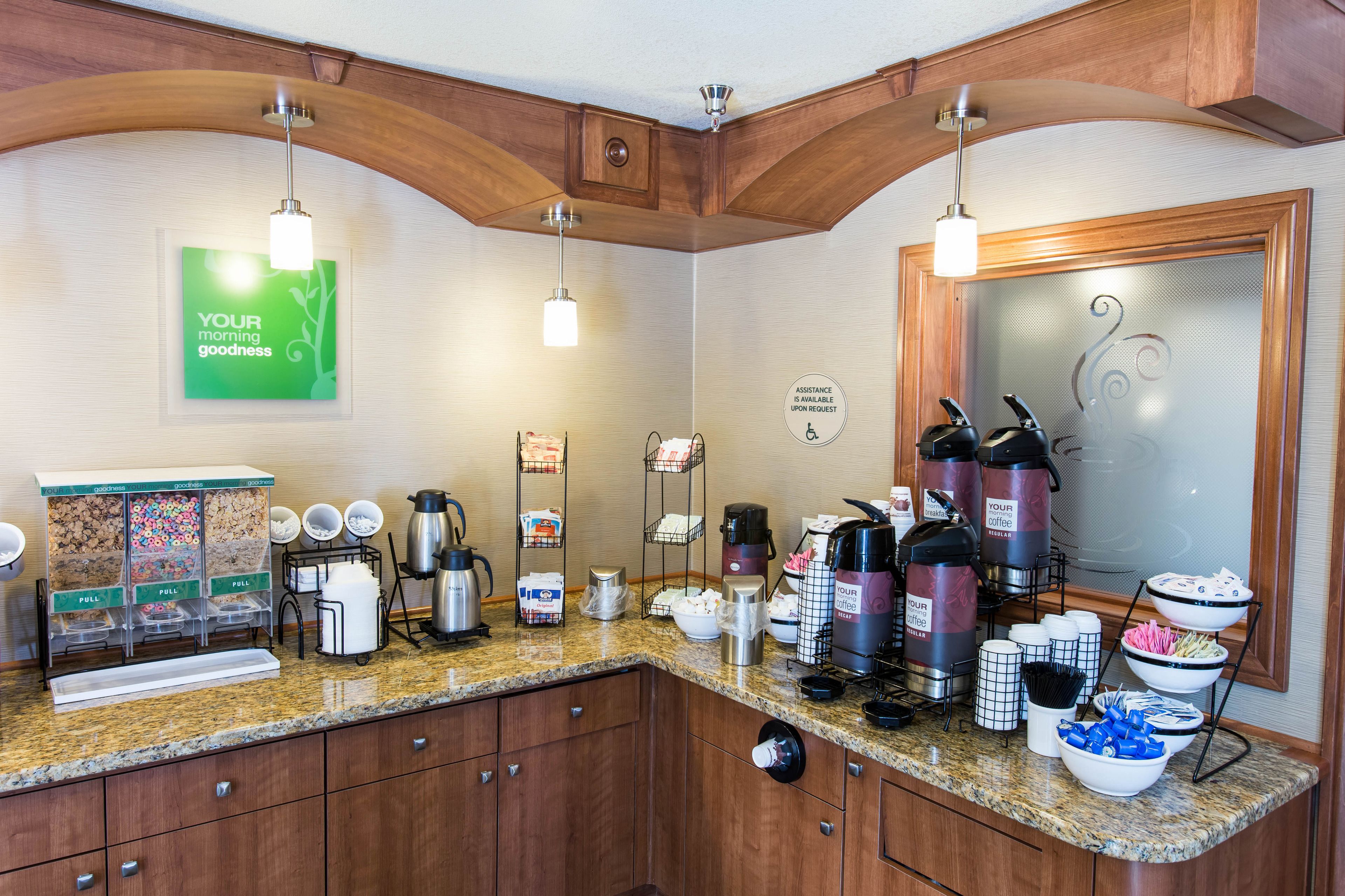 Een ontbijtruimte in een hotel met een koffiehoek, automaten voor eten en een bord met de tekst 'Geniet van je dag'.