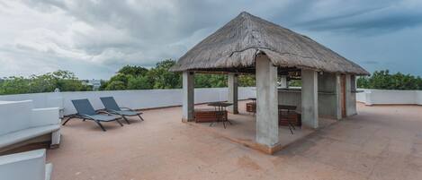 A covered outdoor seating area with lounge chairs and a table, surrounded by a low wall and a thatched-roof structure.