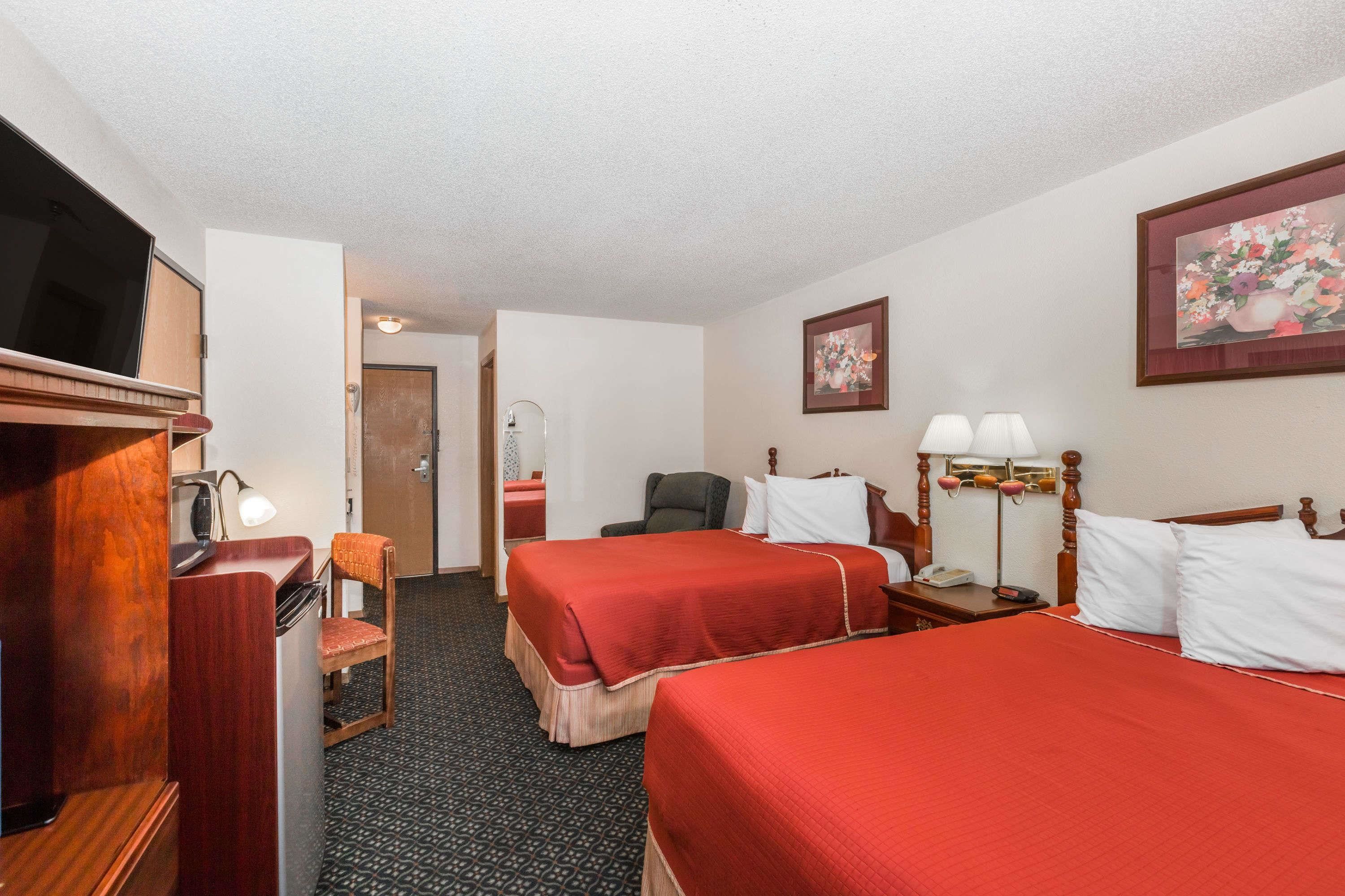 A hotel room with two beds, a desk with a lamp, a chair, a TV, and a painting on the wall.