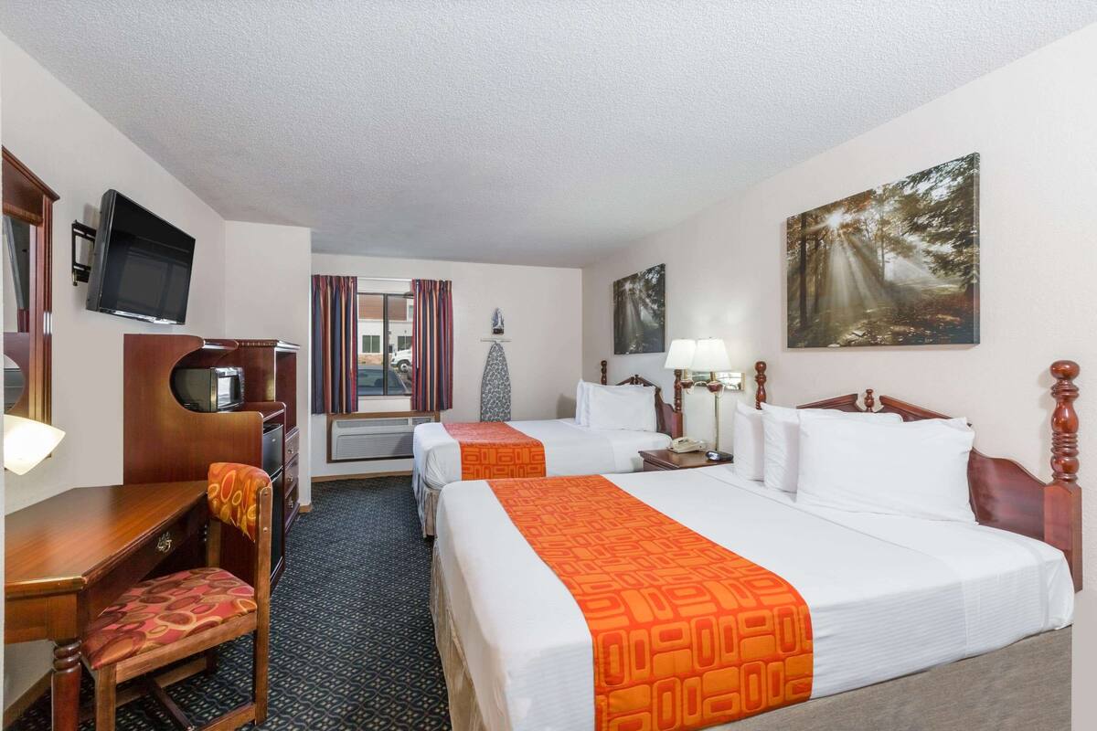 A hotel room with two beds, a desk with a chair, a television, a painting, and a window with curtains.