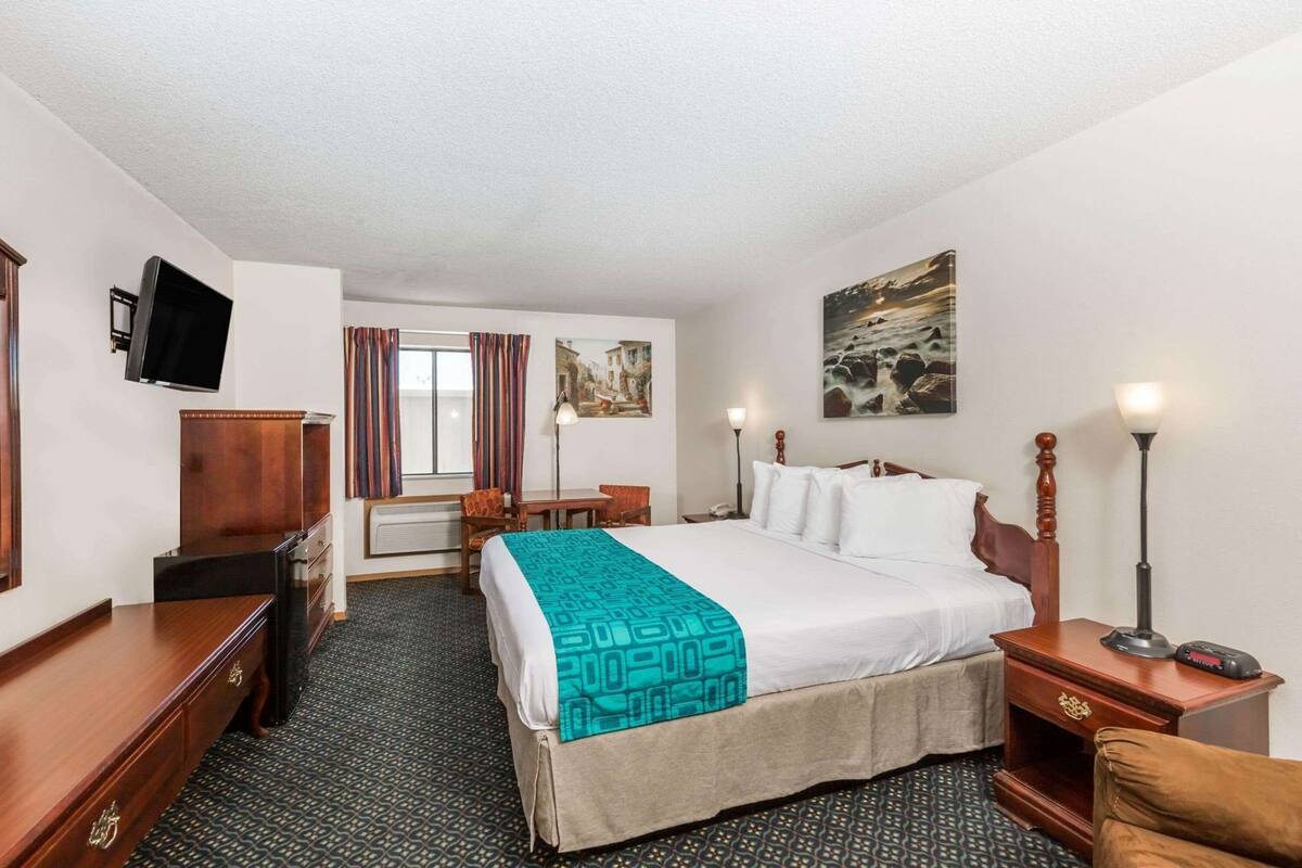 A hotel room with a bed, a desk, a chair, a TV, a lamp, and a painting on the wall.