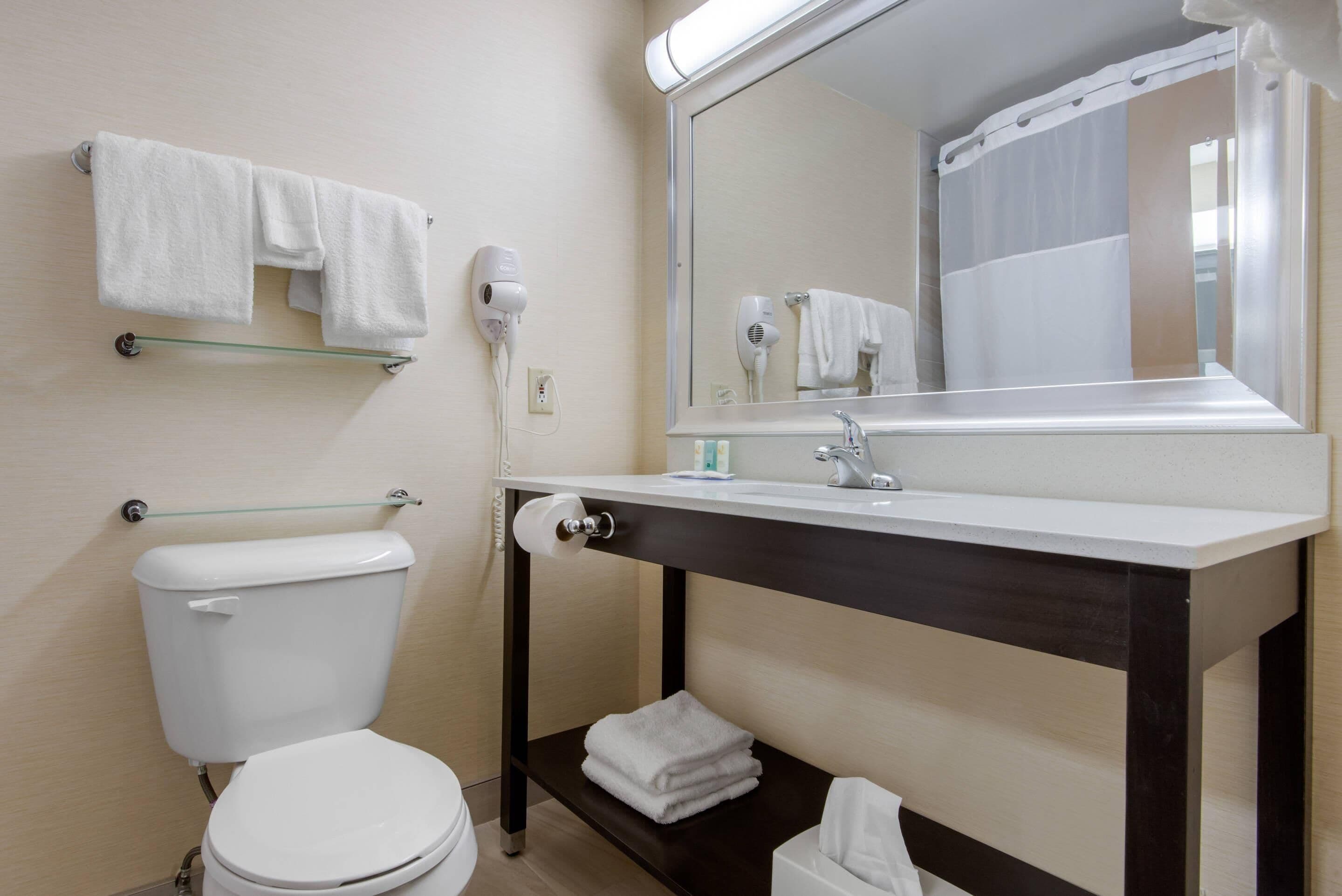 Un baño con inodoro, toallero con toallas, secador de pelo, espejo y lavamanos con dispensador de toallas.