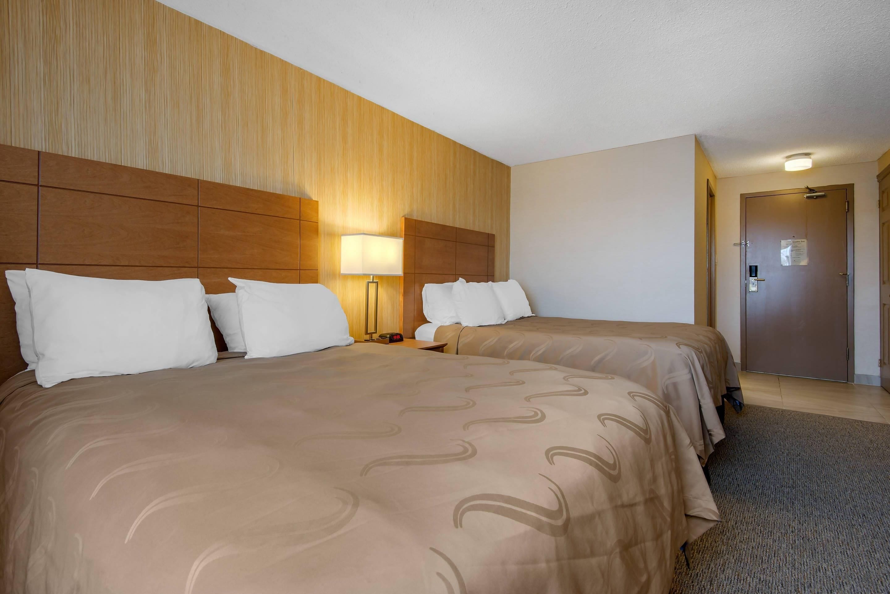 Habitación de hotel con dos camas, cabecero de madera, ropa de cama color crema y lámparas de noche, además de una puerta con cerradura.