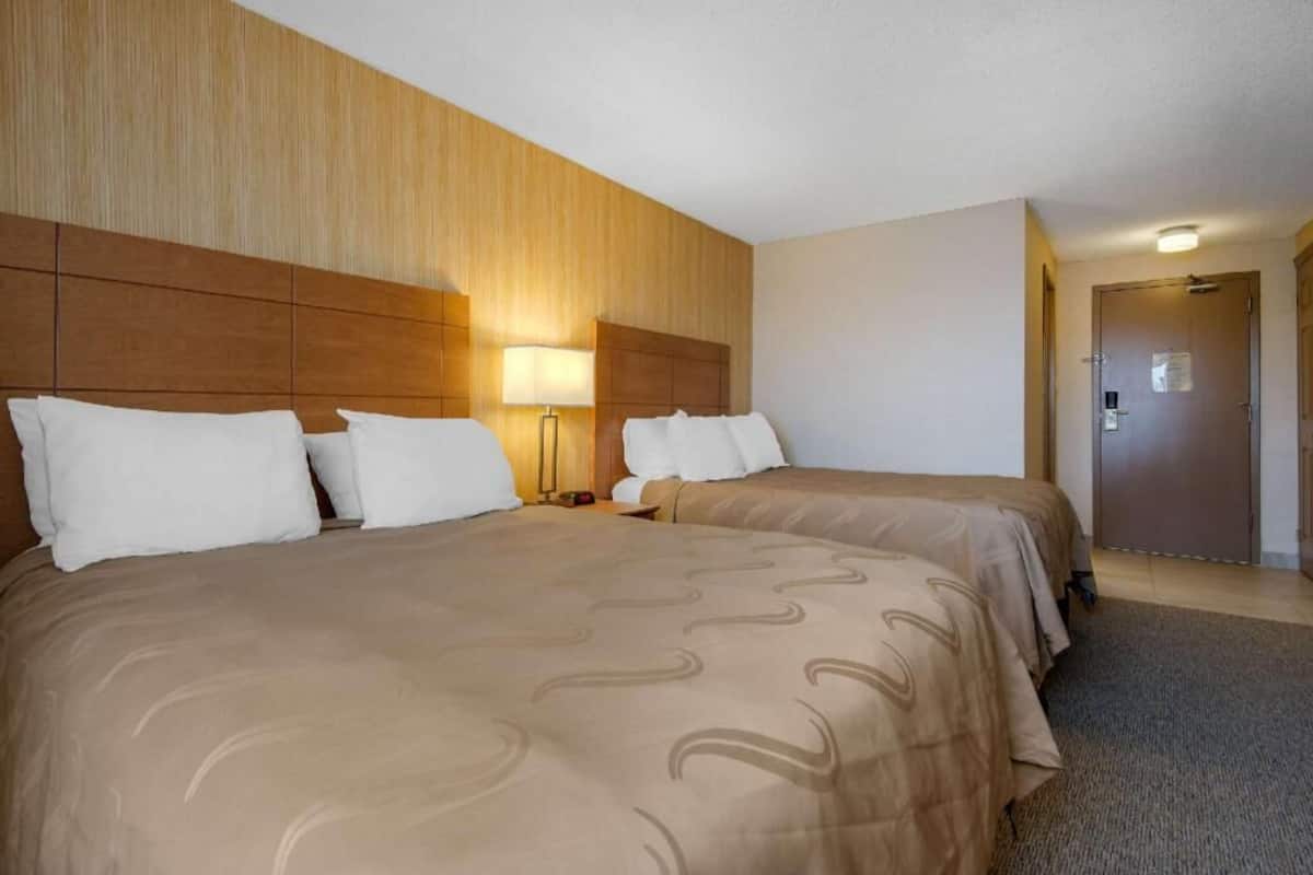 A hotel room with two beds, a wooden headboard, a nightstand with a lamp, and a door.