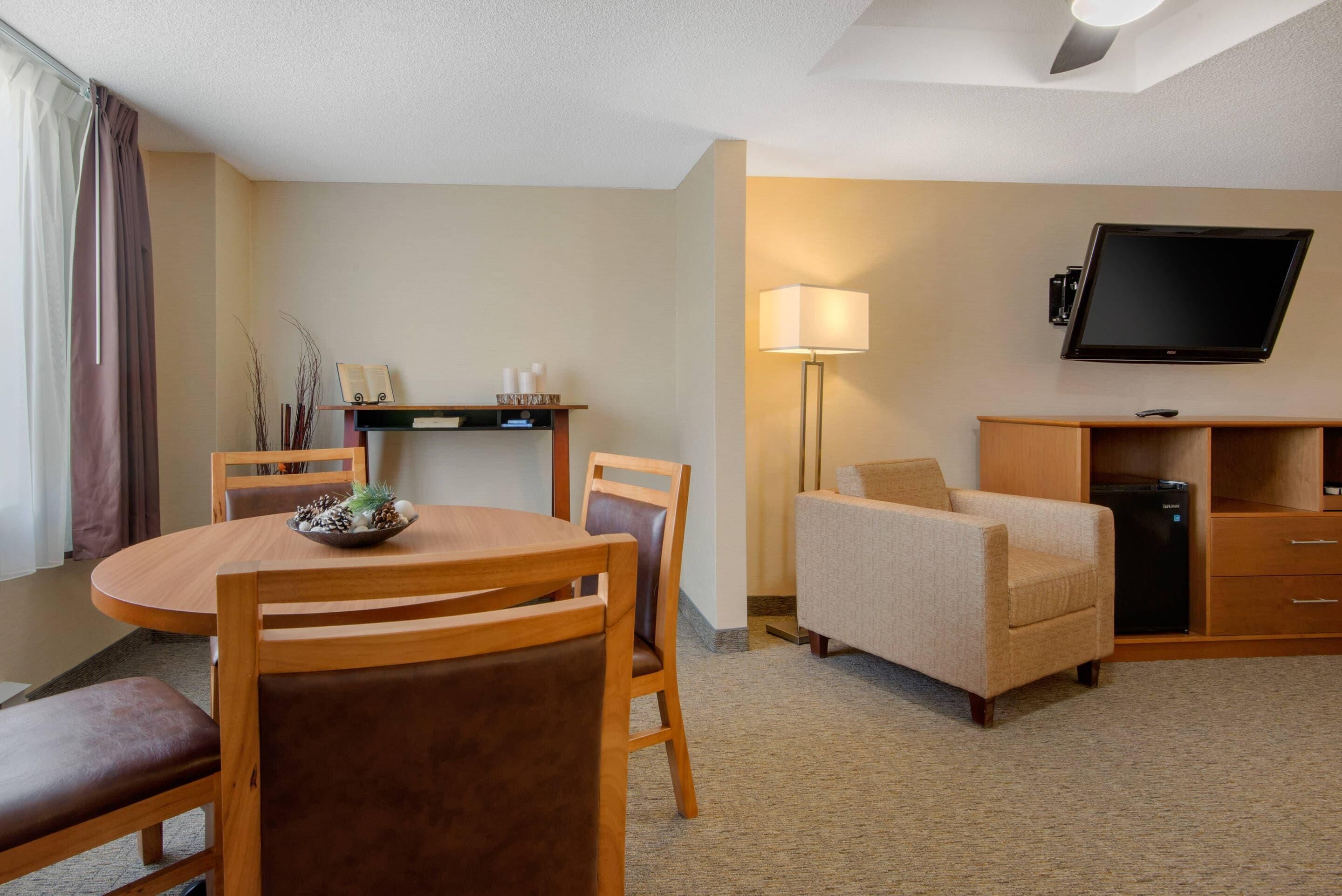 Hotelkamer met een ronde eettafel, houten stoelen, een bank, een aan de muur bevestigde televisie en een klein kastje.